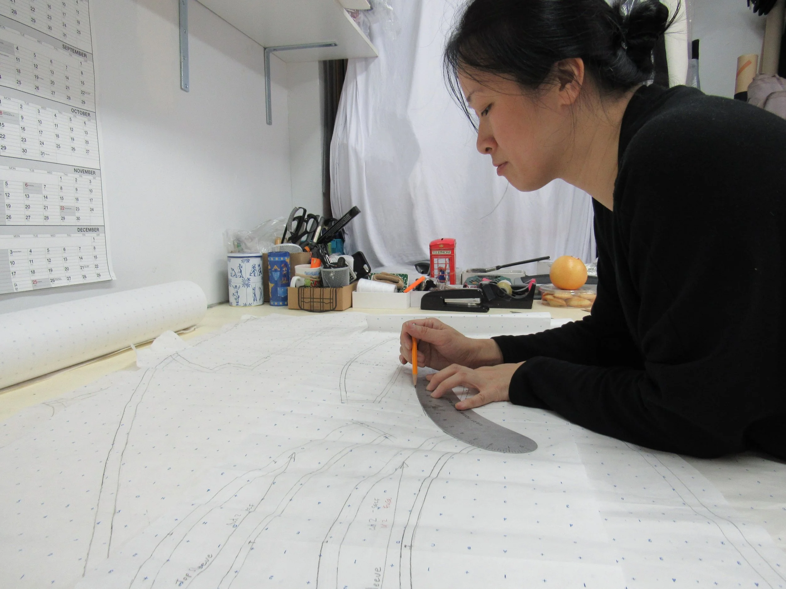 A woman with dark hair styled in a messy bun is leaning over a large sheet of white paper with dotted lines and markings, working on a design or pattern at a workspace. The workspace has various items including scissors, pens, a red telephone booth figurine, and a plate with a fruit. The wall behind her has calendars for September, October, November, and December, and there is a white shelf above.