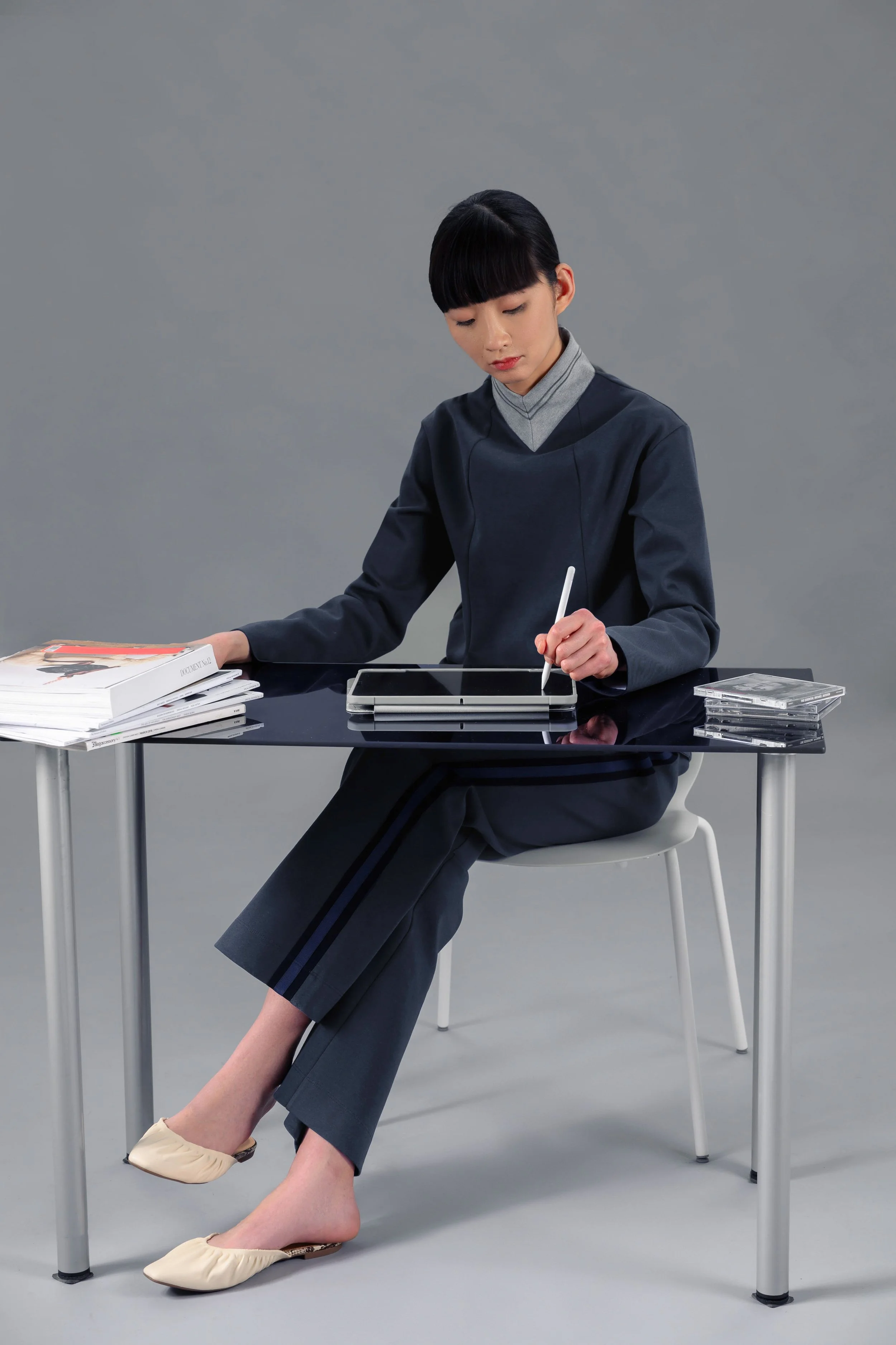 A woman with short black hair and bangs, sitting at a black table against a gray background, wearing a dark gray blazer over a light gray turtleneck, using a stylus on a tablet, with a stack of magazines and a clear plastic cassette case on the table, her feet resting against the table's leg.