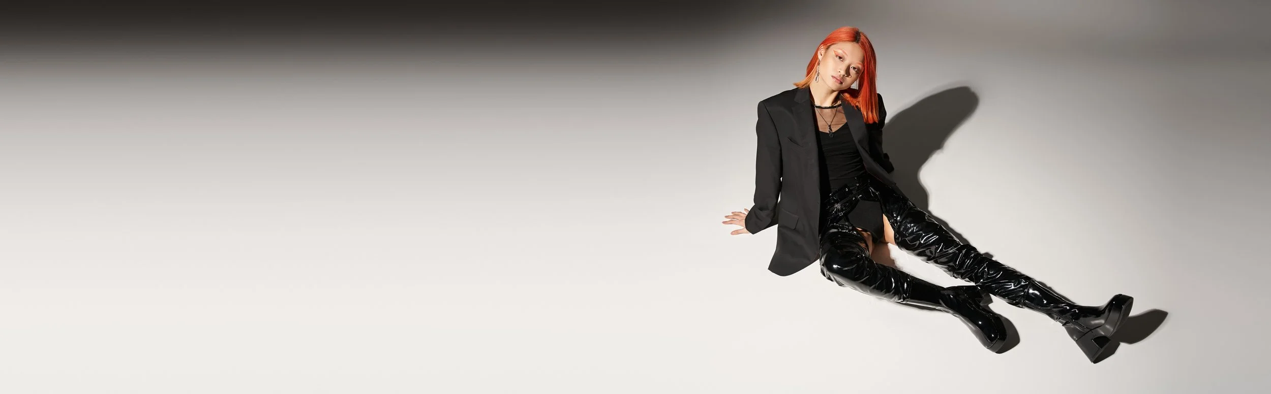 Woman with red hair sitting on a white surface wearing a black blazer, black top, and shiny black thigh-high boots