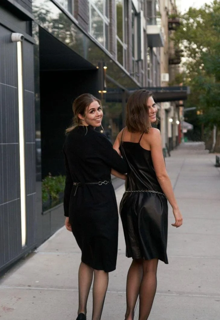 Two women walking on a city sidewalk, smiling and looking back at the camera, dressed in black outfits, with modern buildings and trees in the background.