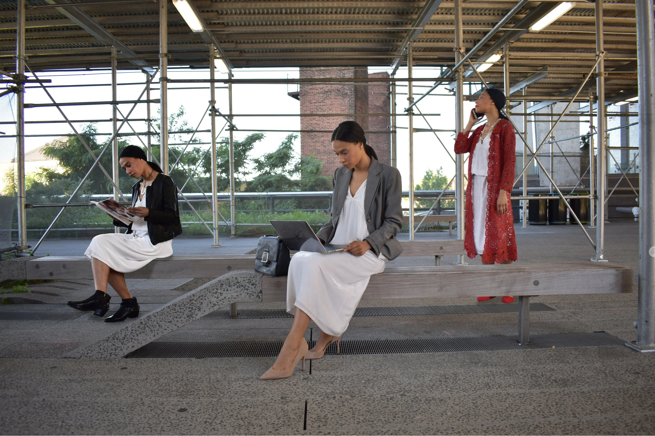 Three women sitting and standing outdoors on a bench near a construction site with scaffolding. One woman is reading a magazine, another is using a laptop, and the third is talking on a cell phone.