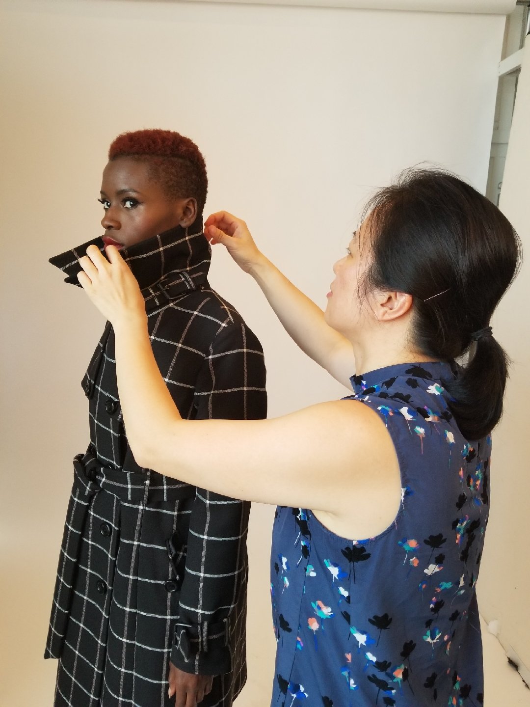 A woman with dark hair is adjusting a checkered trench coat on a woman with short reddish hair.