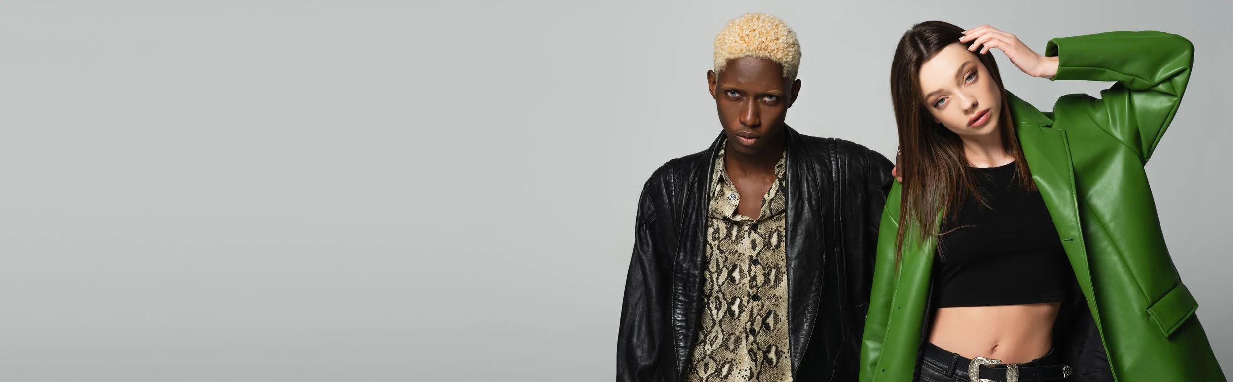 Two fashionable young women posing against a plain gray background. The woman on the left has dark skin, short bleached blonde hair, and is wearing a black leather jacket over a snake print shirt. The woman on the right has light skin, long brown hair, and is wearing a bright green leather jacket over a black crop top, with her hand touching her head.