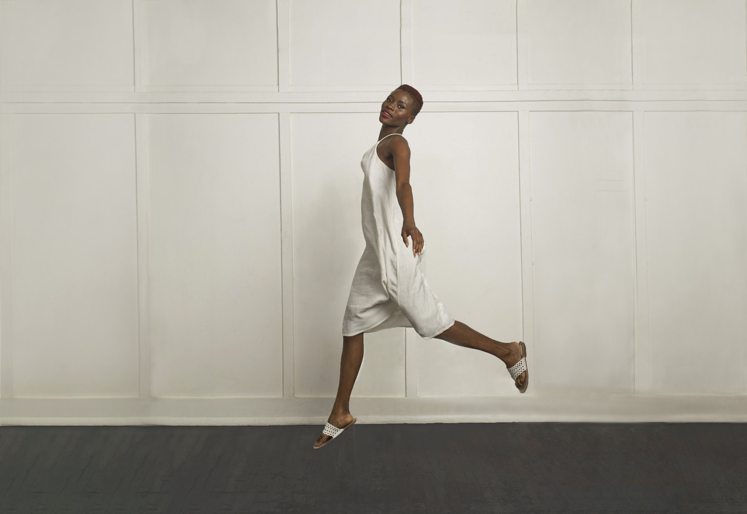 A woman with dark skin and short red hair poses against a white paneled wall. She is wearing a silky, white slip dress and open-toed sandals, balancing on her left foot with her right leg extended backward and her right arm extended downward.