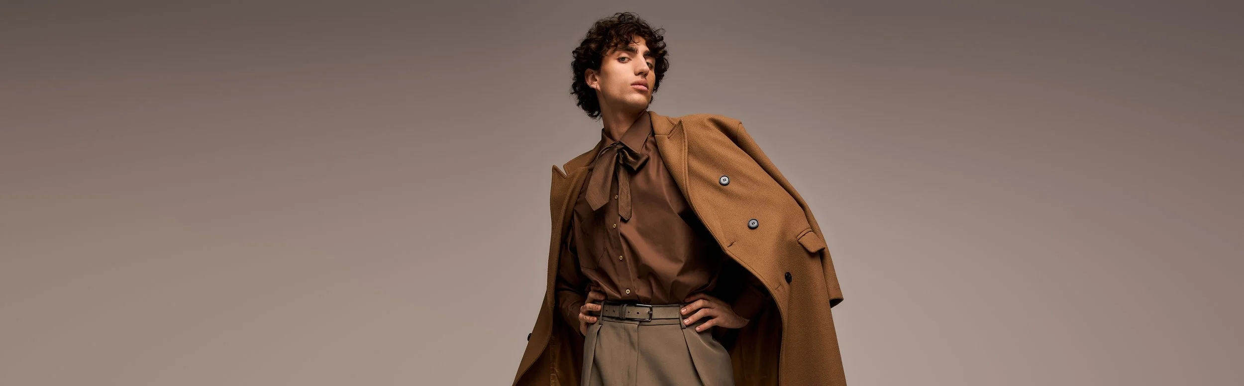 A woman with short curly hair wearing a brown button-up shirt, khaki high-waisted trousers, and a tan coat draped over her shoulder, posing against a plain neutral background.