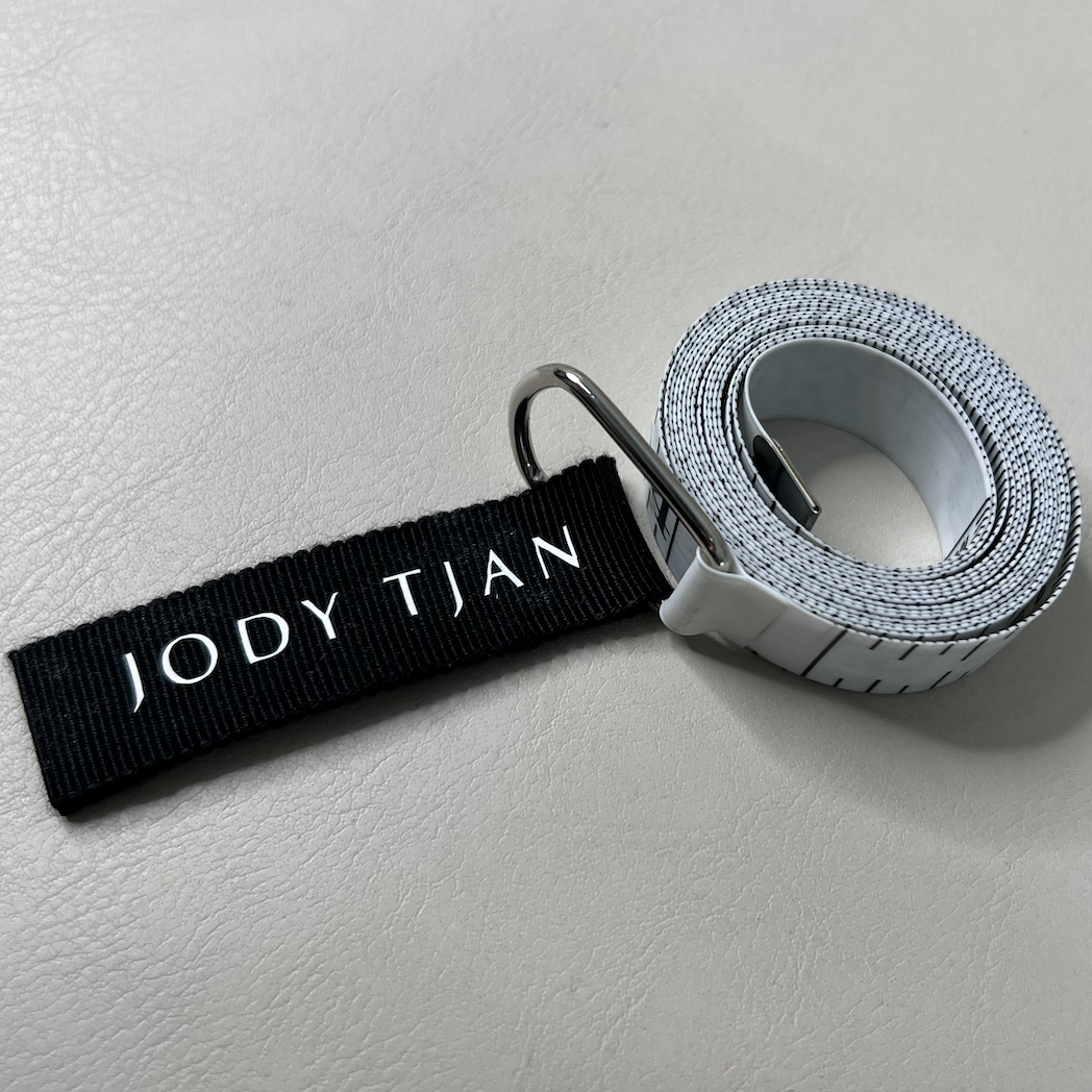 A rolled-up white golf belt with black stripe details and a metal clasp, attached to a black strap labeled 'JODY TJAN' in white lettering.