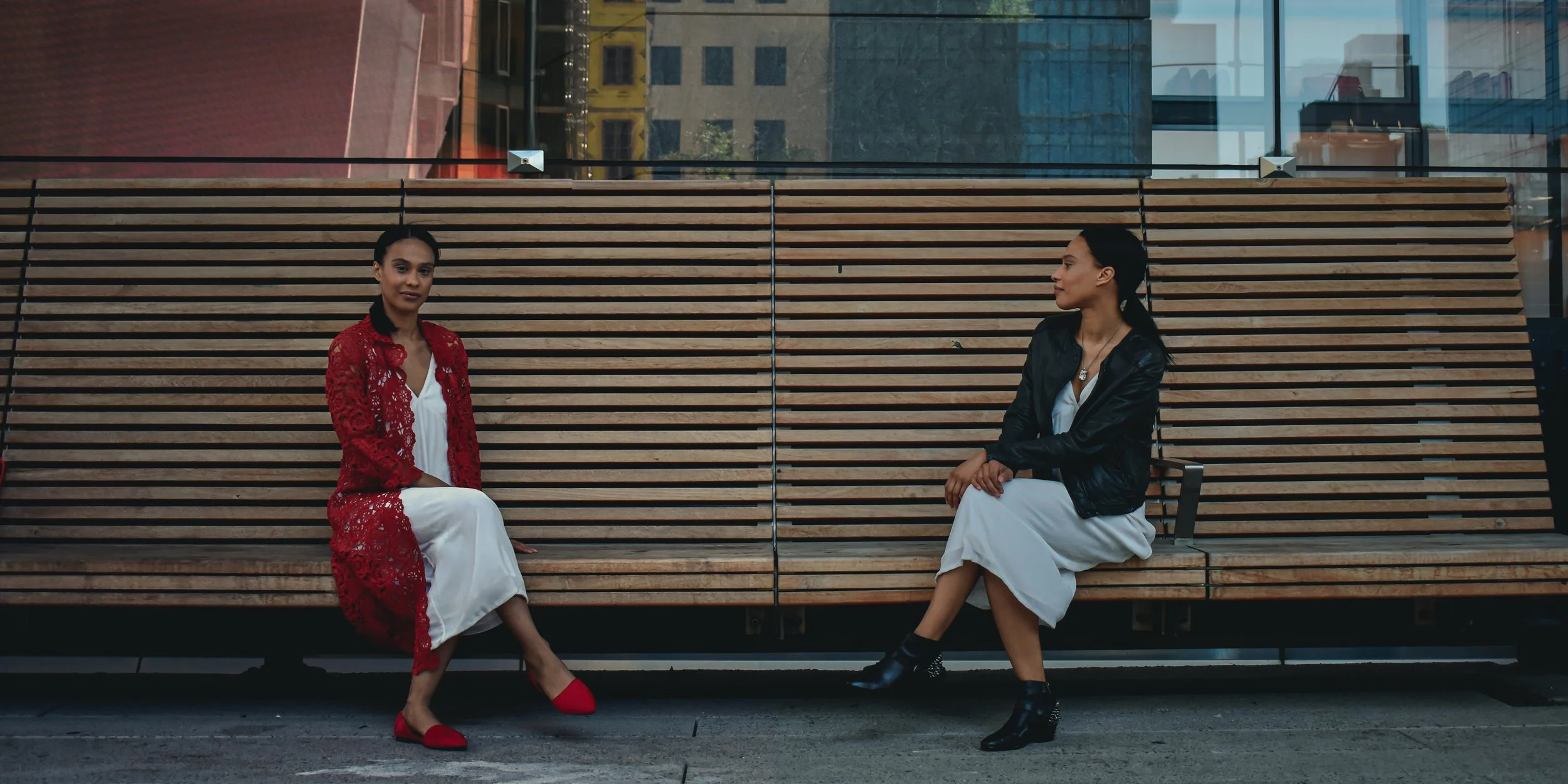 Two women sitting on a wooden bench outdoors in an urban setting, one wearing a red lace jacket and white dress, the other wearing a black leather jacket and white dress, with glass buildings in the background.