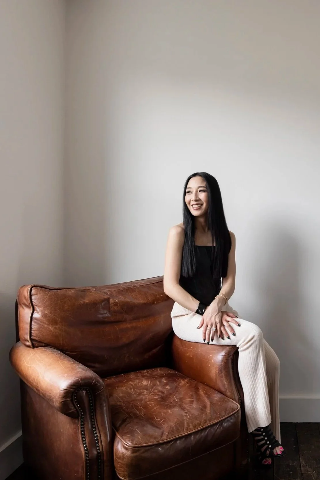 A woman with long black hair, wearing a black sleeveless top and cream-colored pants, sitting on a brown leather sofa against a plain white wall, smiling and looking to her left.