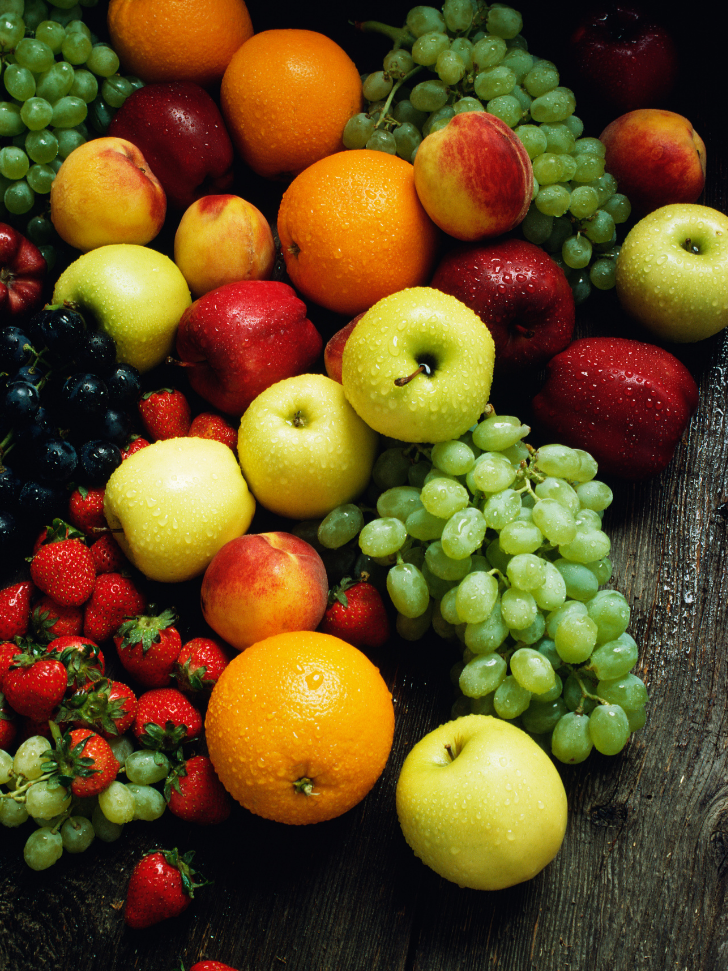 An assortment of fresh fruits including green and red grapes, strawberries, apples, peaches, and oranges on a dark wooden surface.