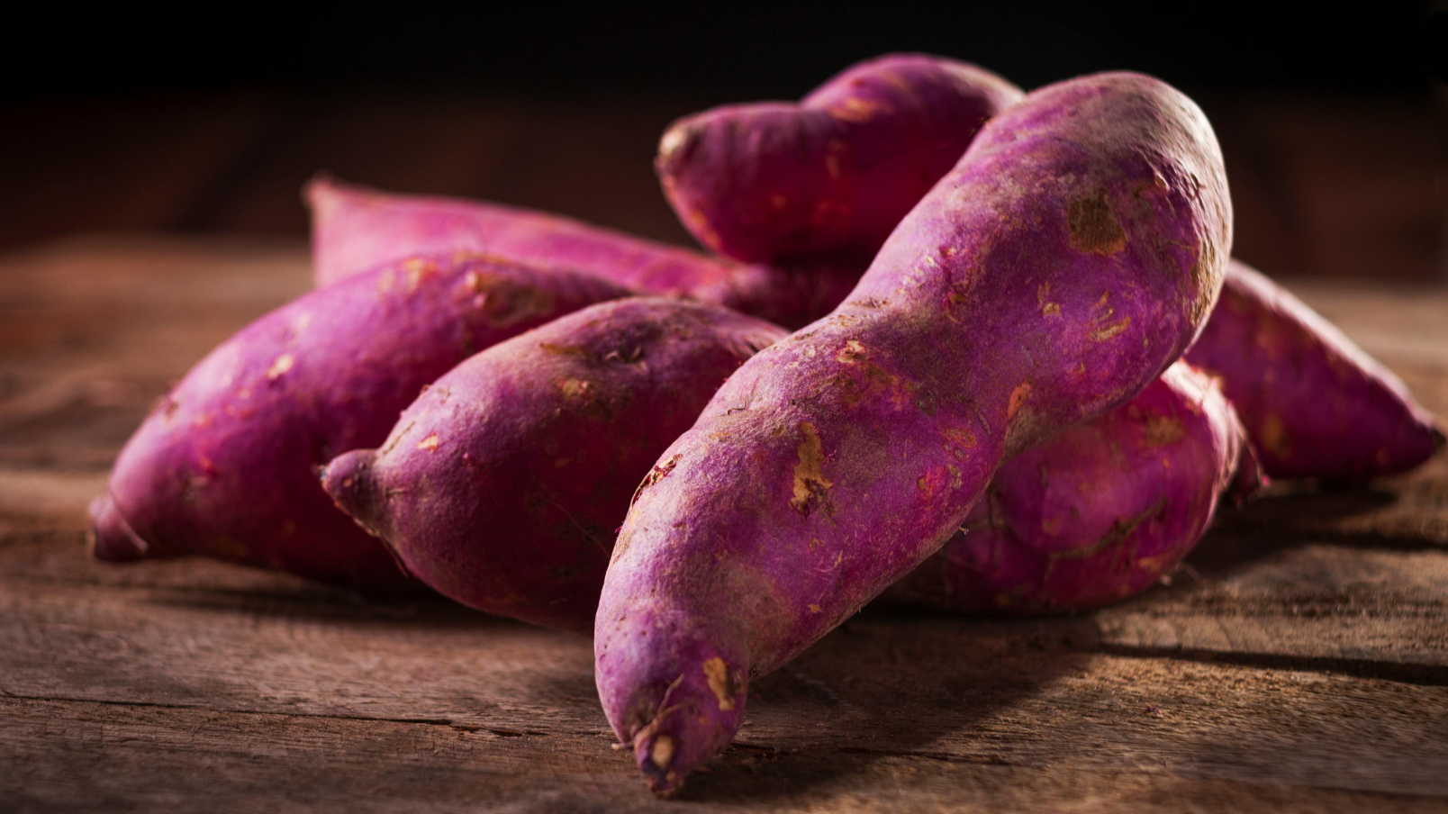 Several purple sweet potatoes on a wooden surface, with a dark background.
