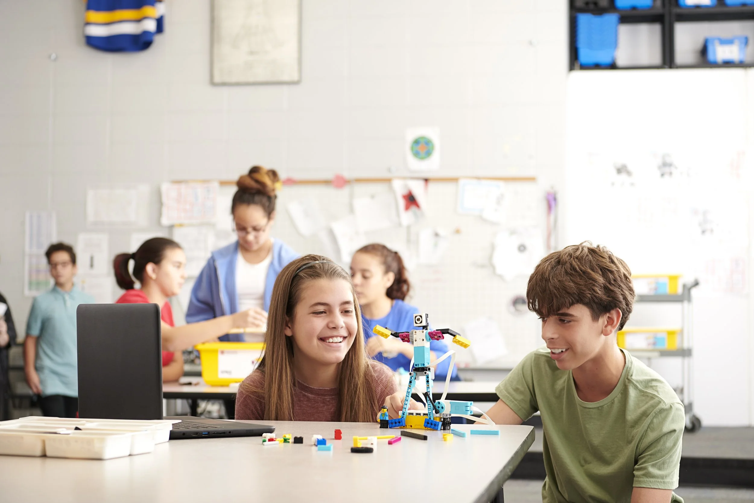 Twee kinderen maken een robot van LEGO in een klaslokaal, terwijl andere kinderen op de achtergrond bezig zijn.