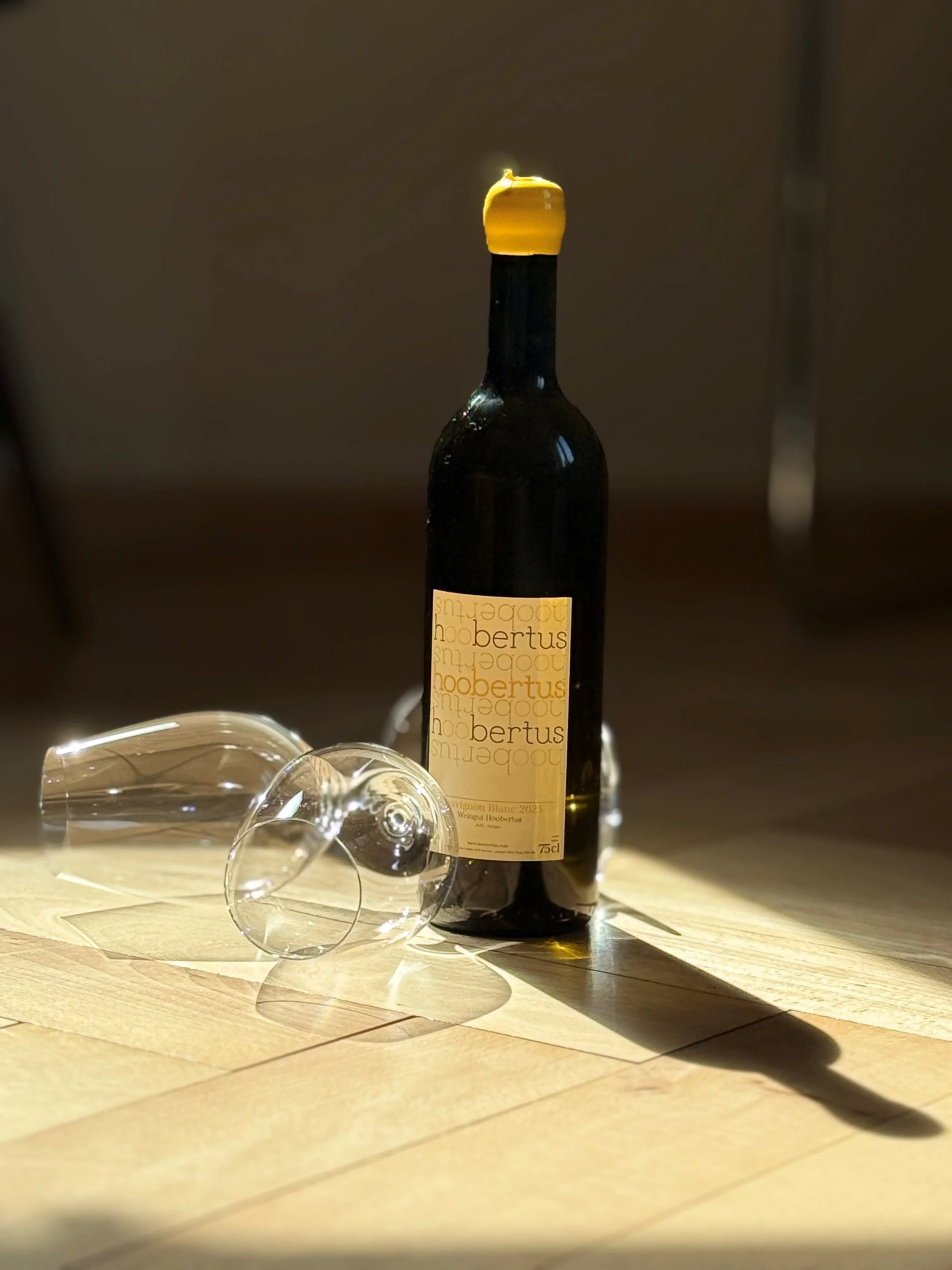 Eine Flasche Wein mit einem gelben Korken und zwei leeren Weingläser liegen auf einem Holztisch. Das Licht fällt schräg, wodurch Schatten entstehen.