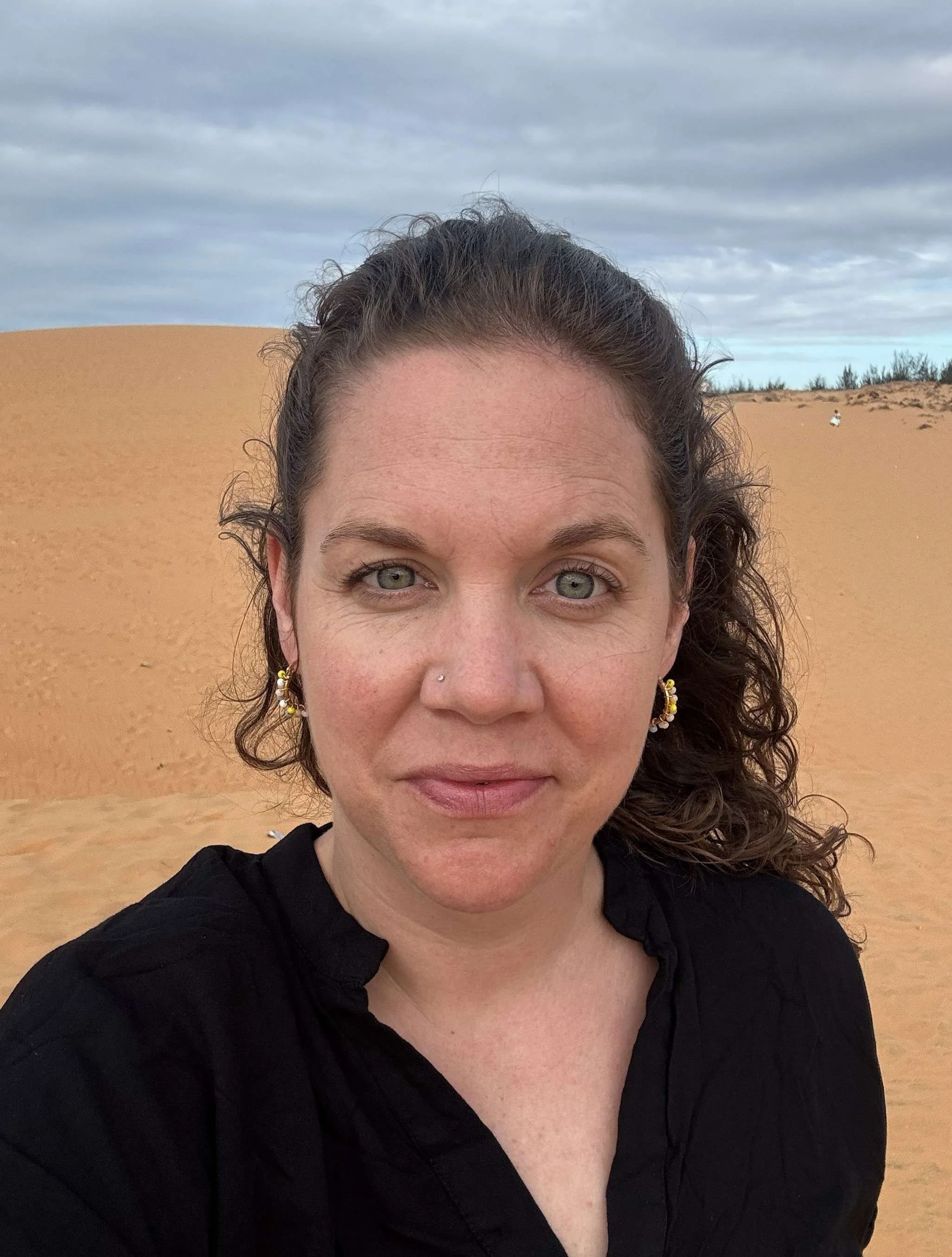 Nahaufnahme der Therapeutin Tanja Wüthrich mit braunen, lockigen Haaren vor einer Sanddüne unter einem bewölkten Himmel.