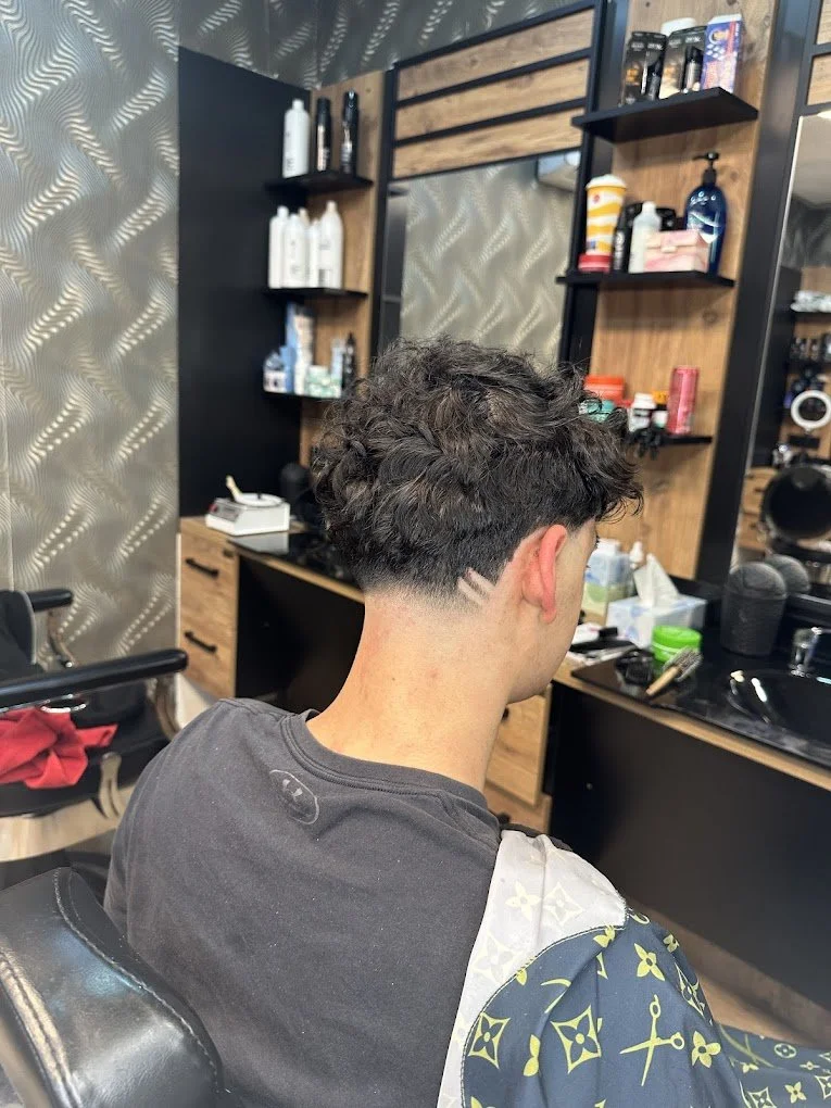 Young man with a haircut, sitting in a barber shop chair with a barber cape. The back of his head shows a shaved design on the right side. The barber shop has shelves with hair products and tools, a large mirror, and a patterned wall in the backgroun