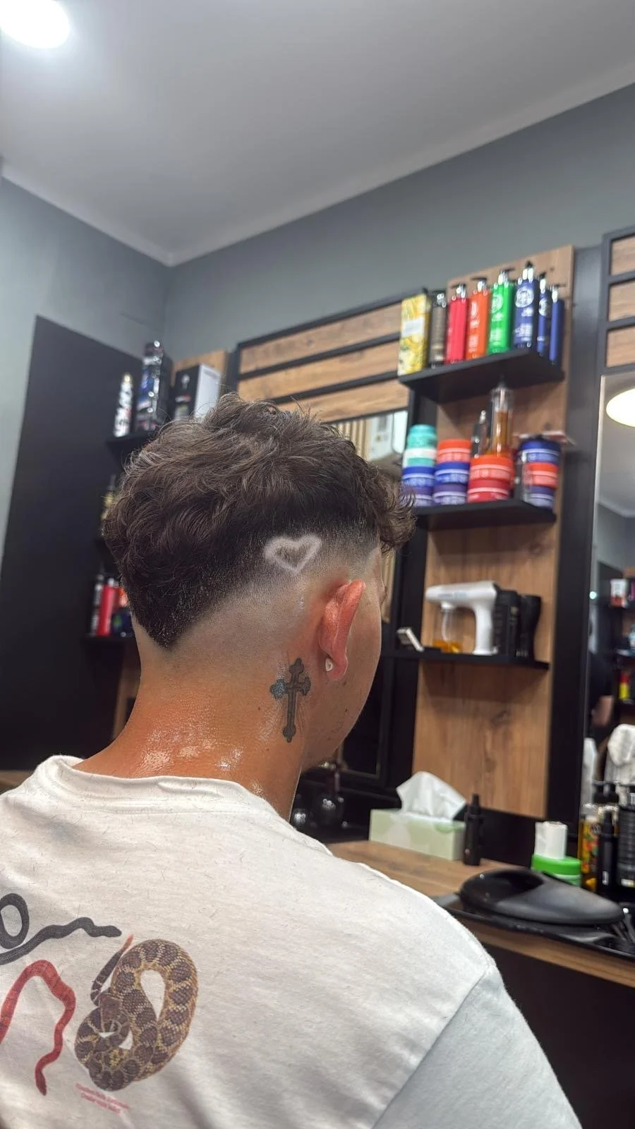Back of a person's head with a short curly hairstyle featuring a heart-shaped design shaved into the side. The person has a tattoo of a cross on the neck and is sitting in a barbershop.