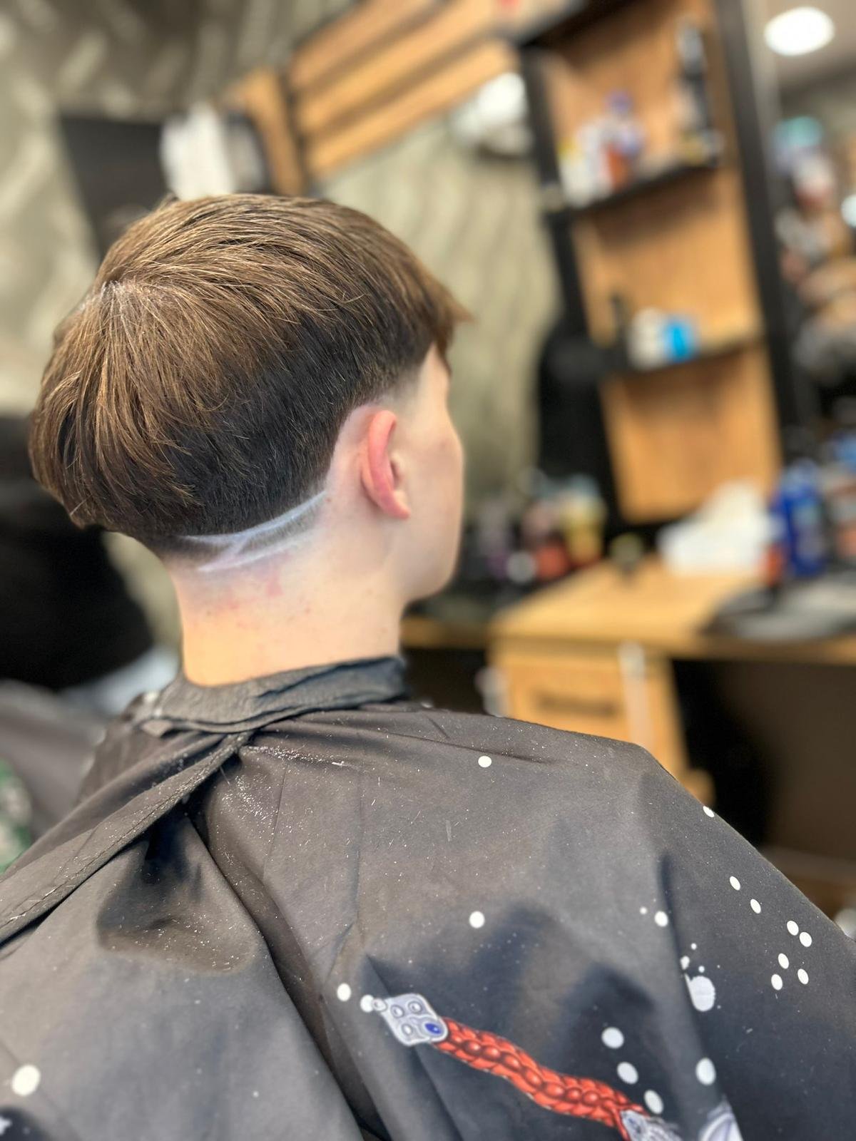 Person with short brown hair sitting in a barber chair, wearing a black barber cape with white paint splatters, inside a barber shop with blurred shelves and tools in the background.