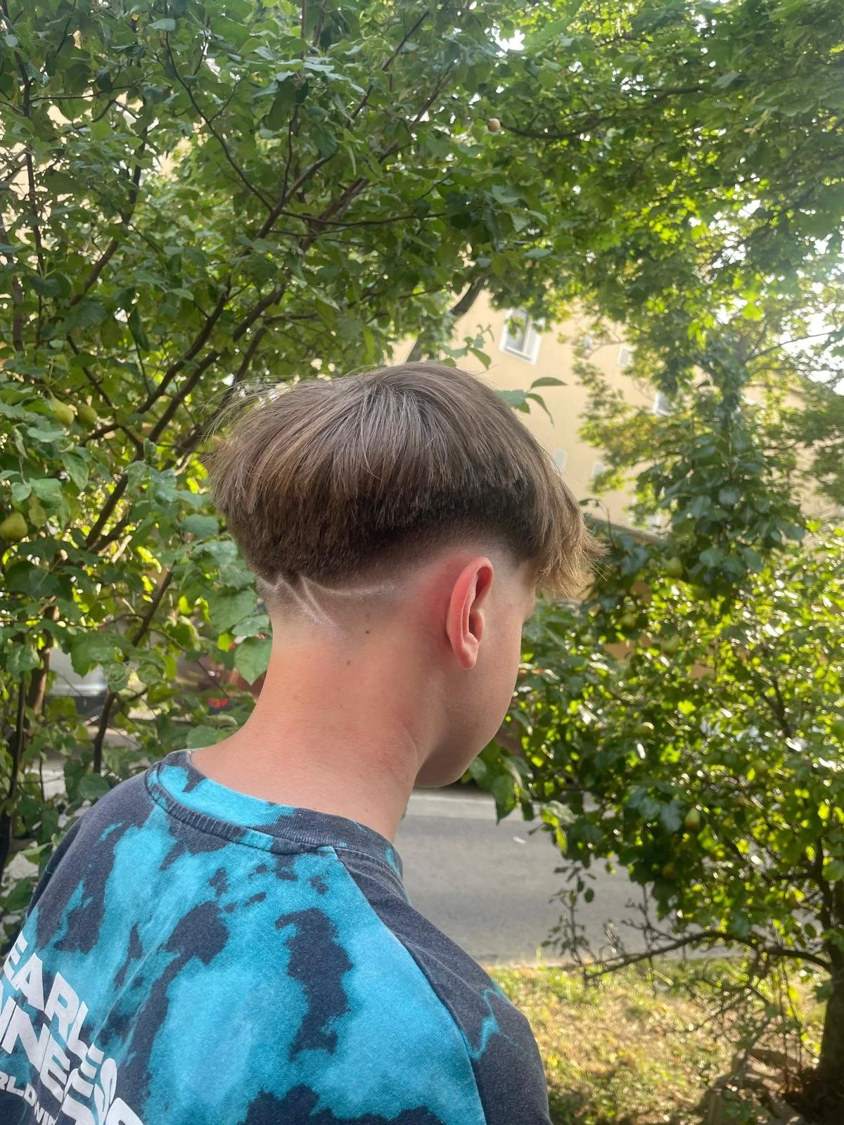 Side view of a young man with a fade haircut, wearing a blue patterned shirt, standing outdoors near leafy green trees and a building in the background.