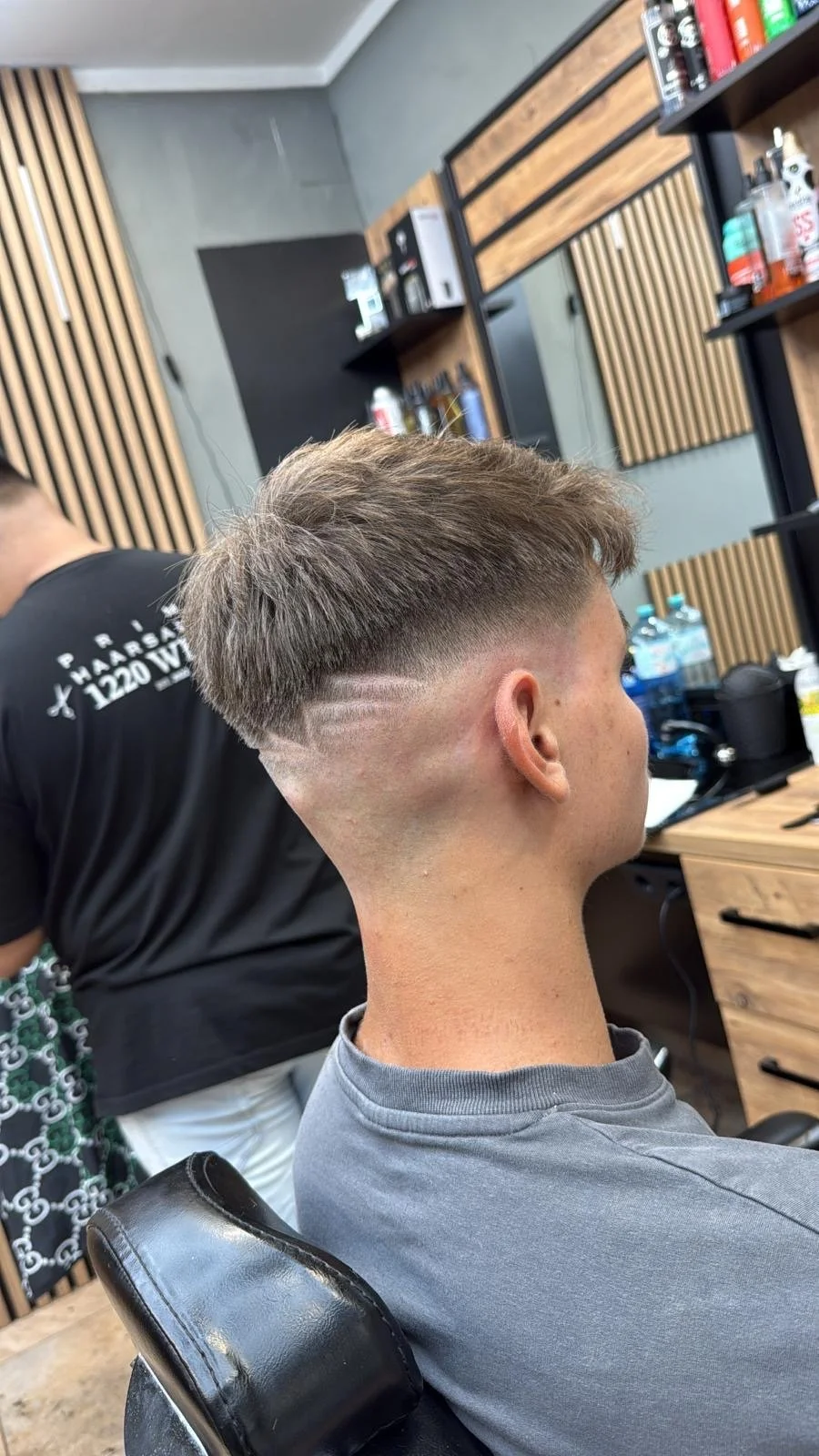 A man in a gray T-shirt sits in a barber shop chair, showing a trendy haircut with blonde, textured hair and a faded undercut with a decorative line shaved into the side. The barber and barber shop interior with wooden panels and hair products are vi