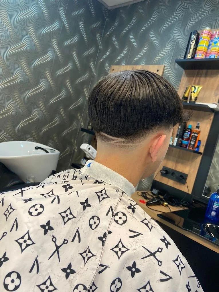 Back of a man's head showing a fresh haircut with a fade and a parallel line design. The man wears a white barber cape with black Louis Vuitton pattern. The barbershop has textured wallpaper, a sink, shelves with hair products, and barber tools.