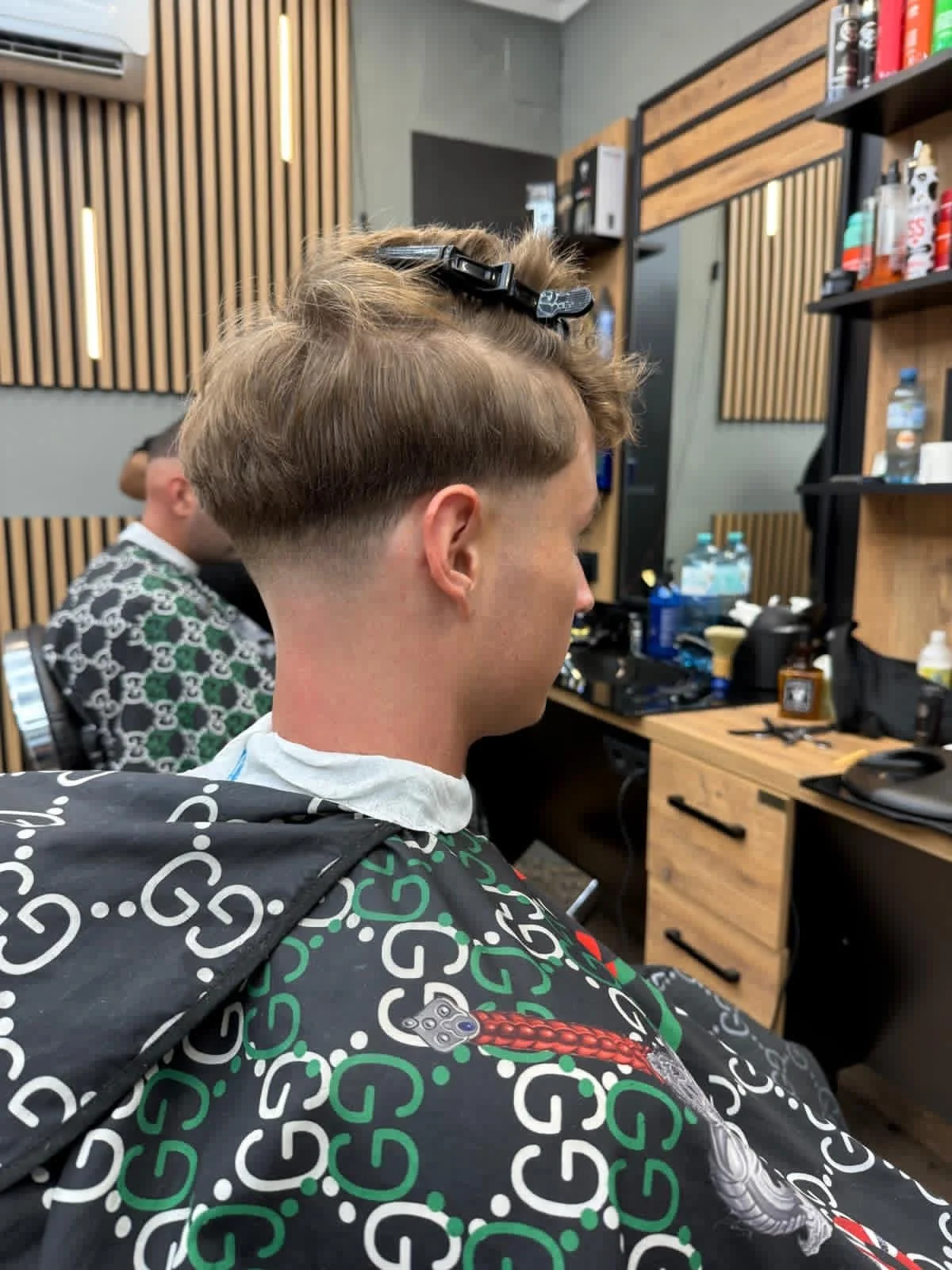 A man getting a haircut in a modern barber shop, seated in a chair with a cape, with another person in the background also receiving a haircut. The barber shop has wooden and black accents with shelves and tools on a counter.