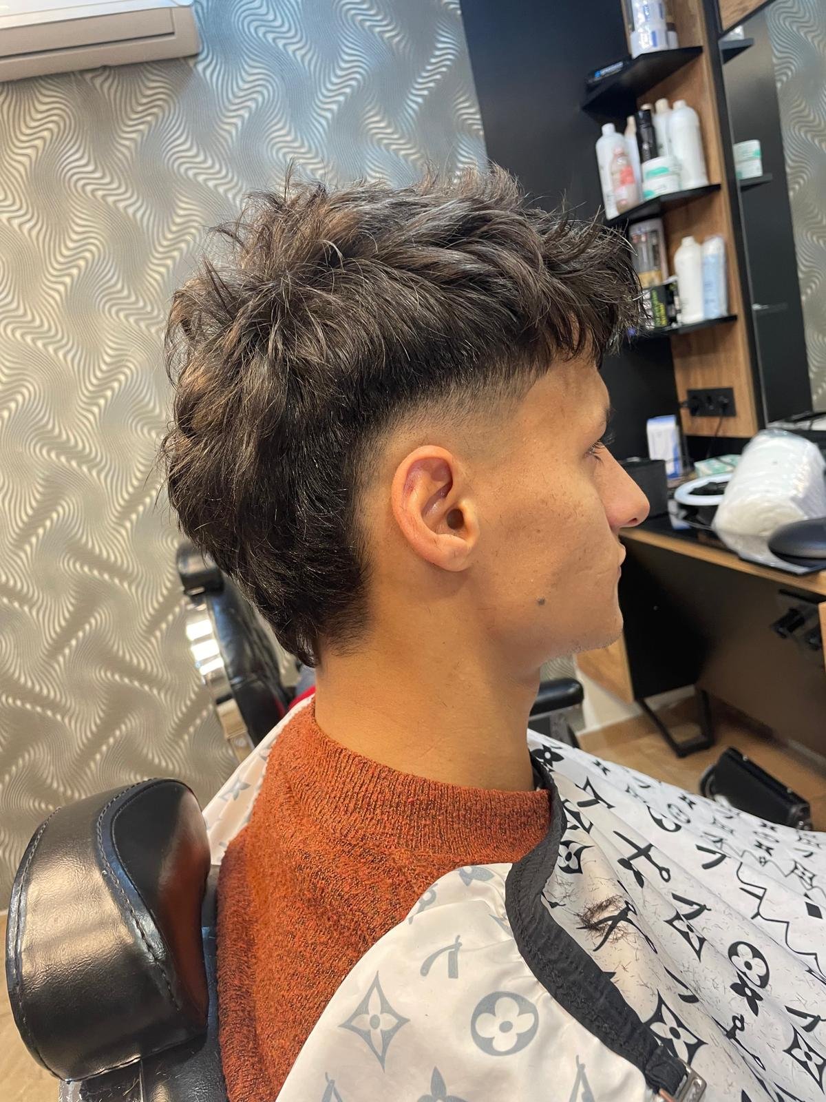 A man sitting in a barber's chair after getting a haircut, with a side fade and textured top. He is wearing a barber cape and an orange sweater.