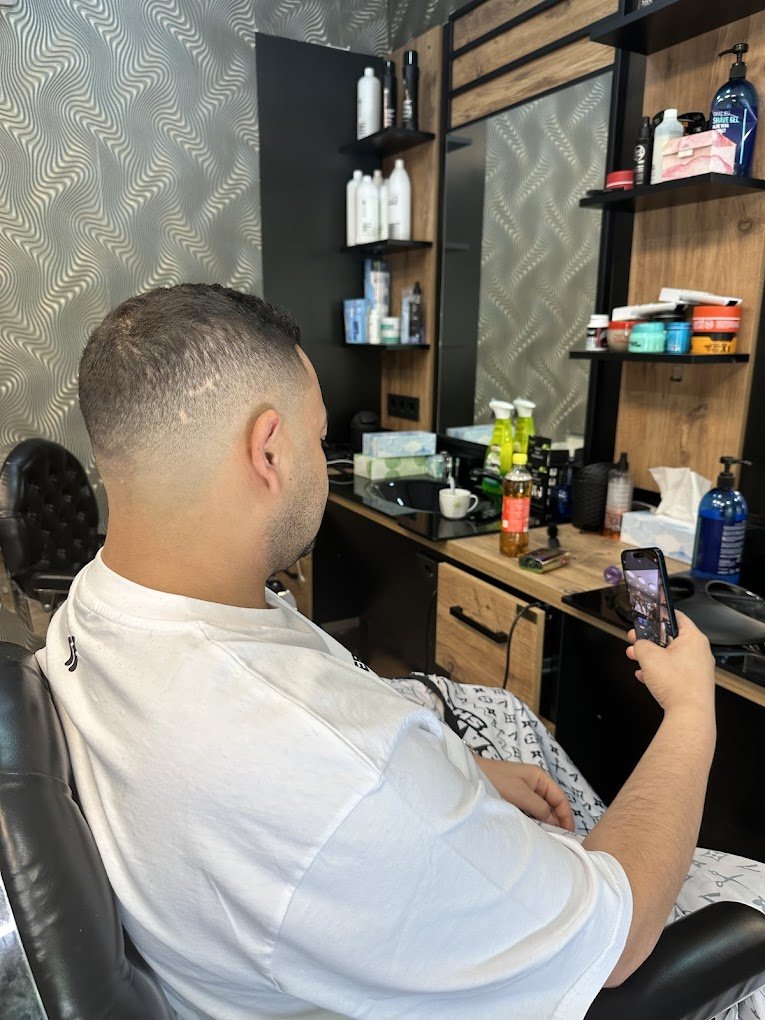 Man with a fade haircut sitting in a barber chair, using a smartphone in a barber shop.