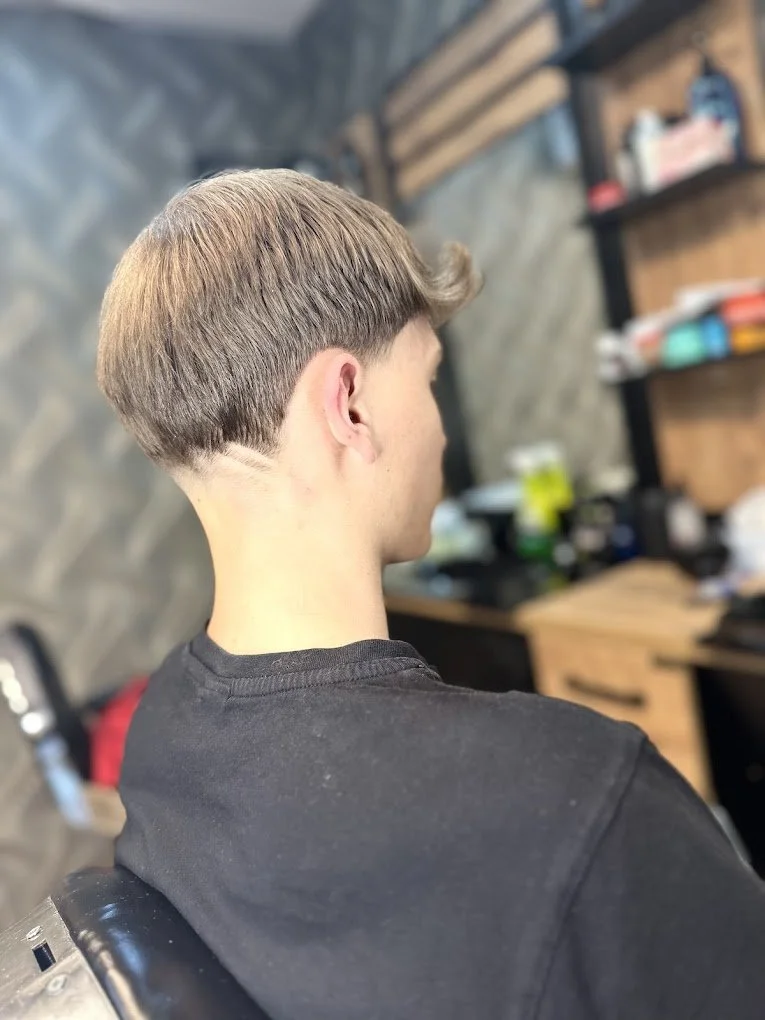 A young man with a short, layered haircut sitting in a hair salon or barbershop, facing away from the camera.