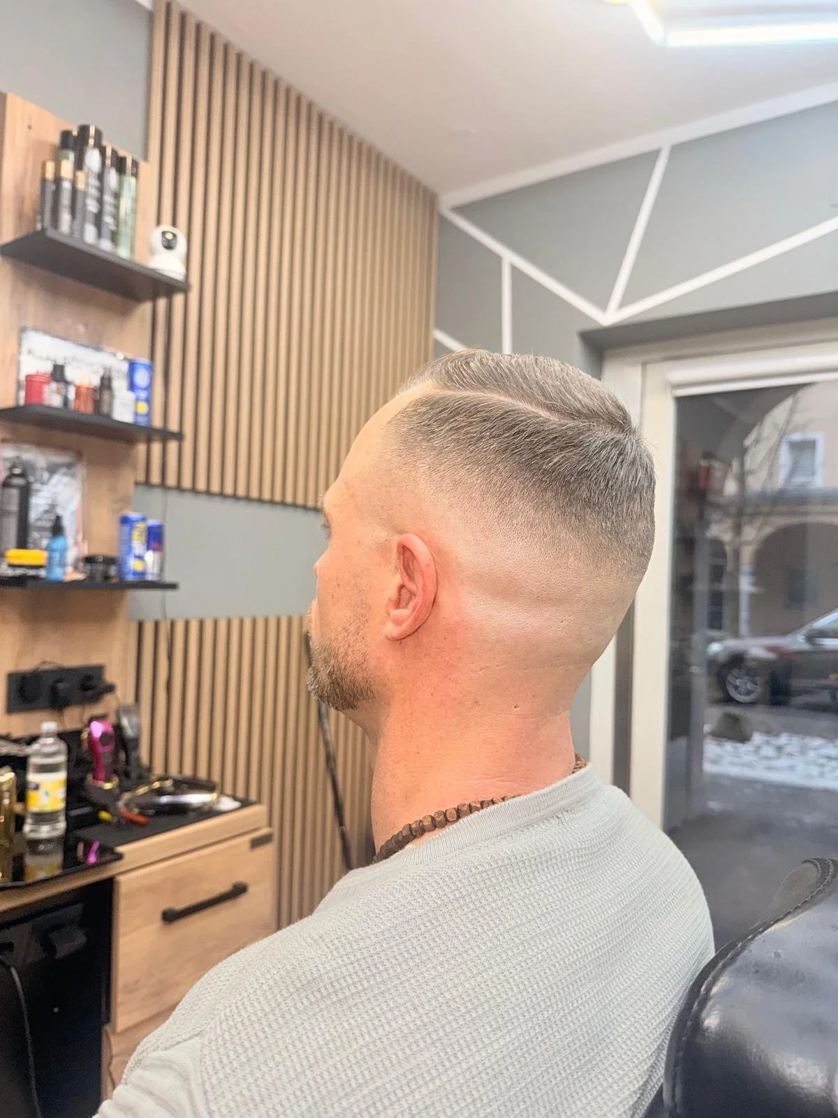 A man with a freshly faded haircut sitting in a barbershop, a view of his side and back of his head, with a background of shelves with barber supplies and decorative wood paneling.