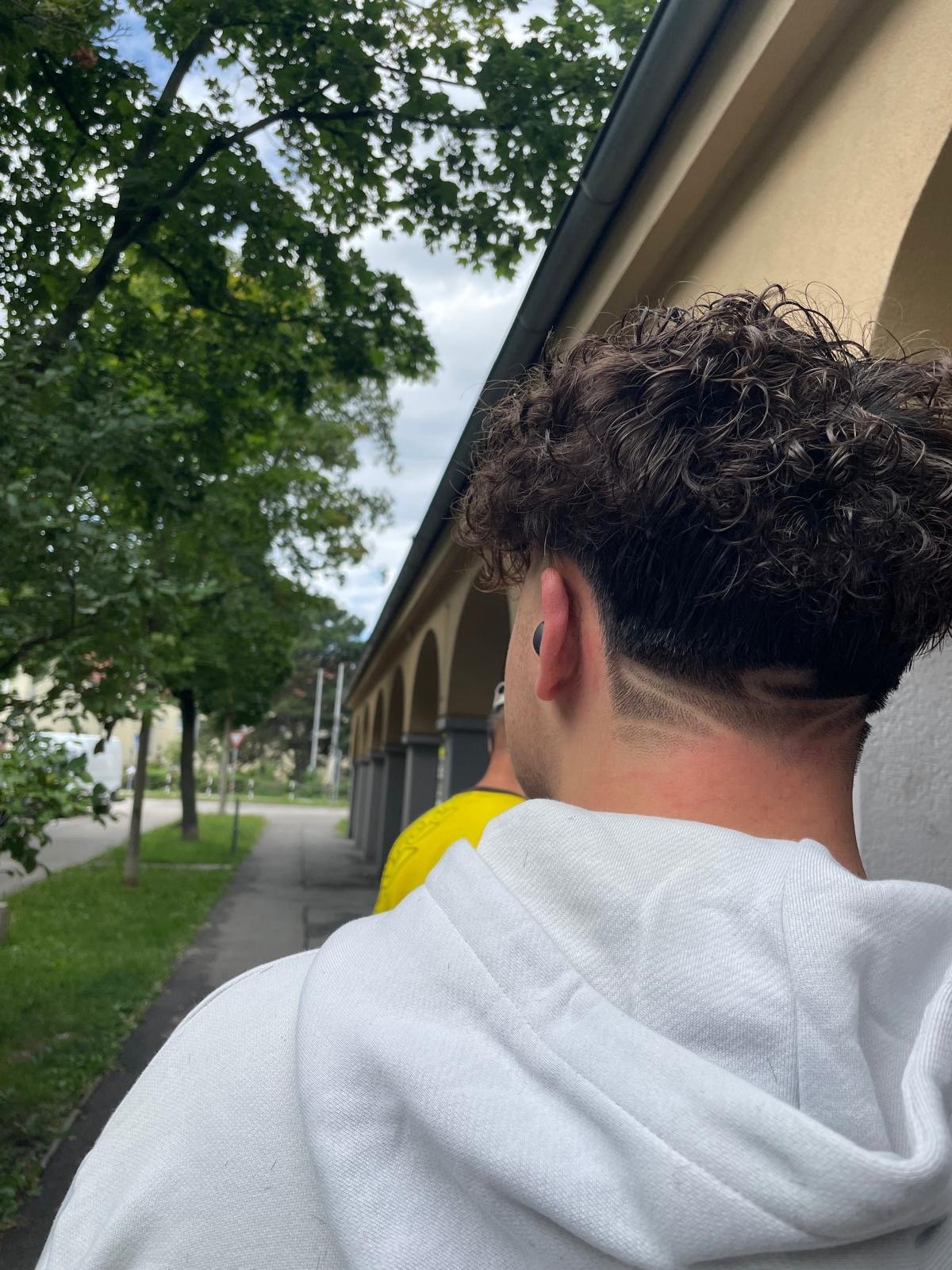Back of young man's head with curly hair and a fade haircut with a line design, standing outdoors on a sidewalk near a building and trees.