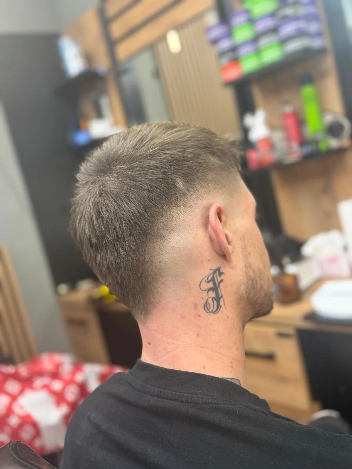A man with a short, faded haircut sitting in a barbershop, showing the left side of his head and neck, which has a tattoo of a cursive letter 'F'.