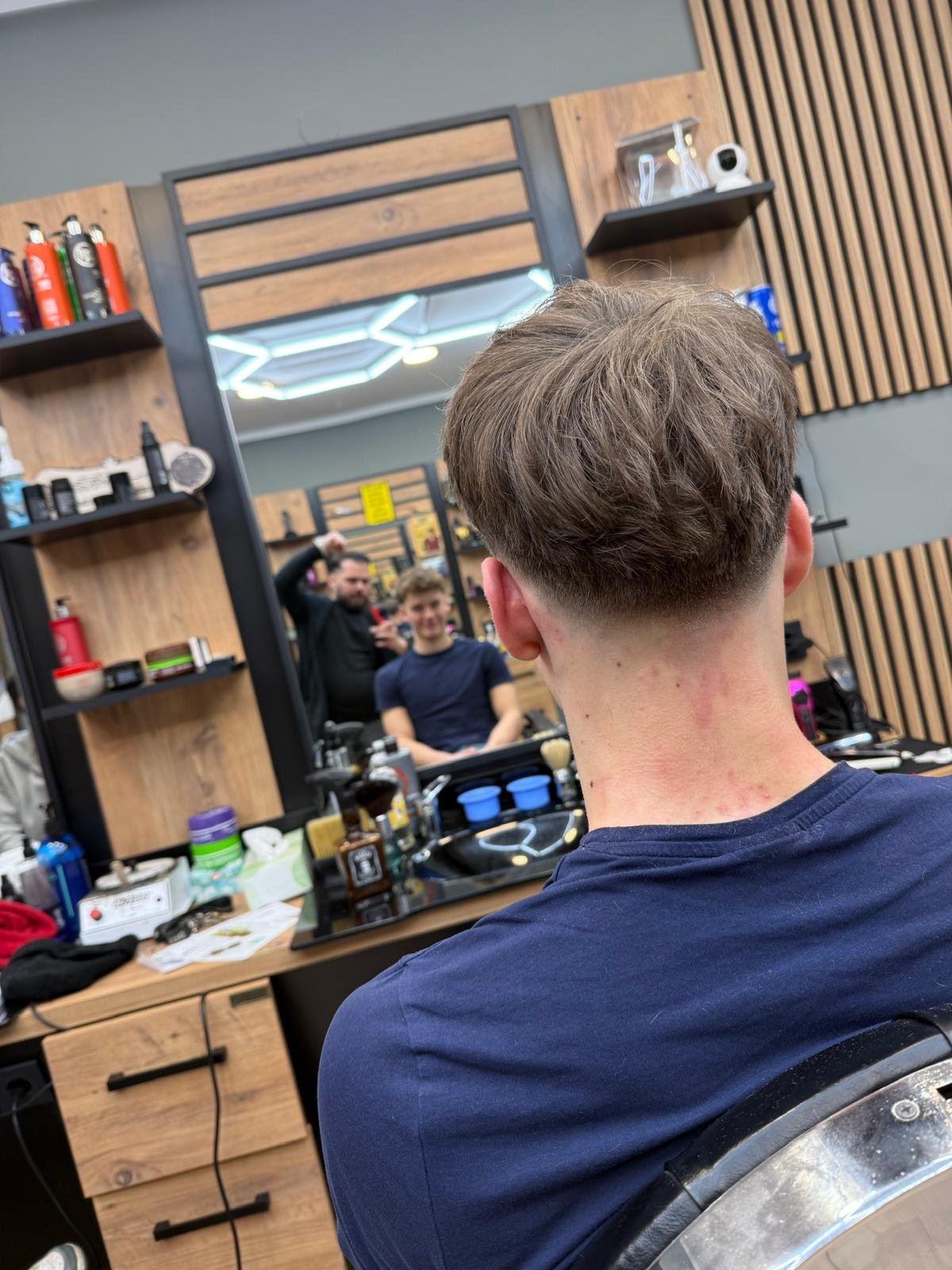 A person with a short haircut sitting in a barber's chair at a barbershop, looking at their reflection in the mirror. A barber and another customer are visible in the reflection, with shelves of grooming products behind them.
