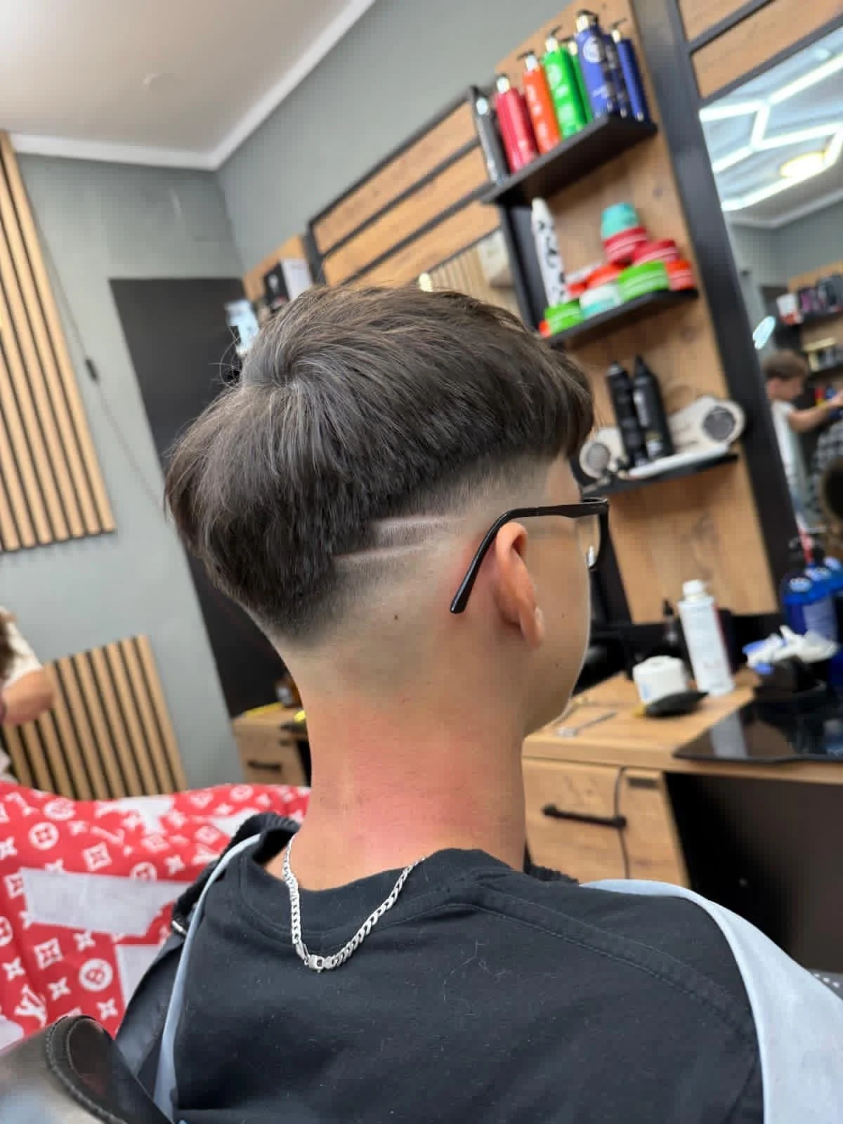 A person with a modern haircut sitting in a barbershop, with shelves of hair products and tools visible in the background.
