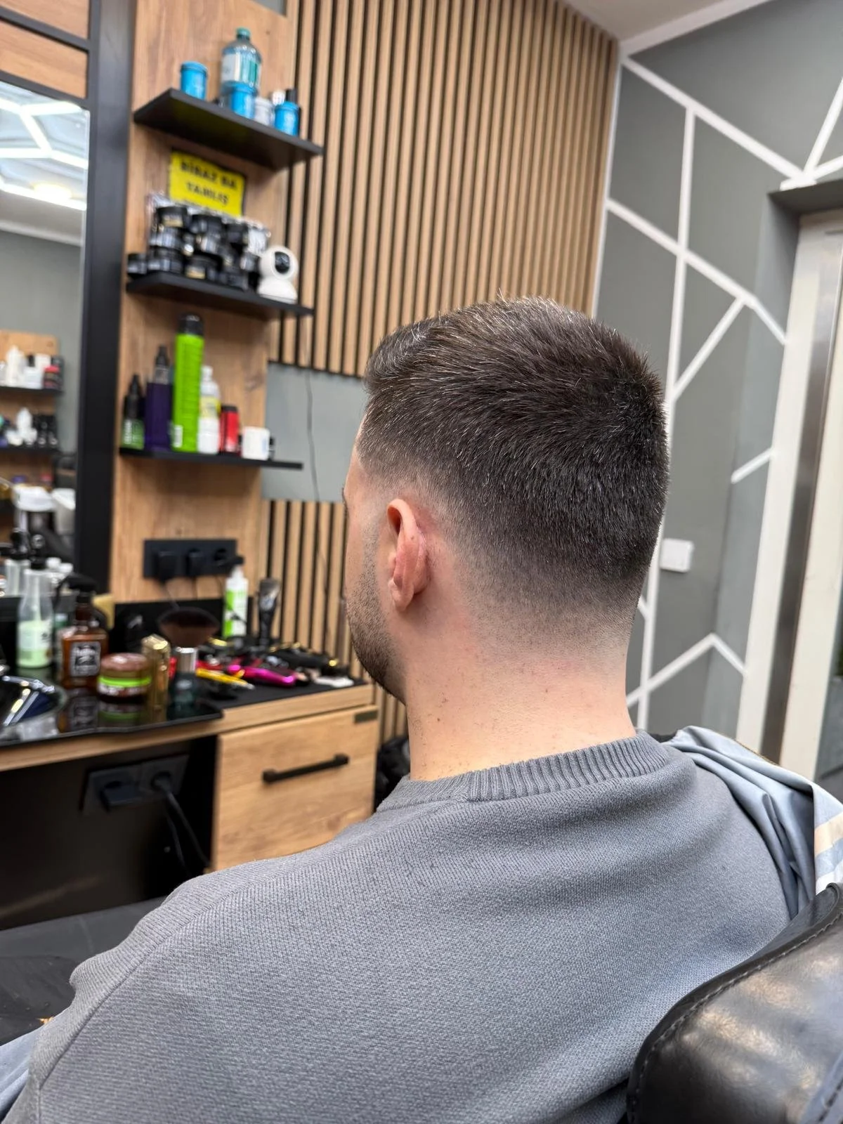 A man with a fresh fade haircut sitting at a barbershop, facing away from the camera, with hair products, tools, and styling equipment on the counter nearby.