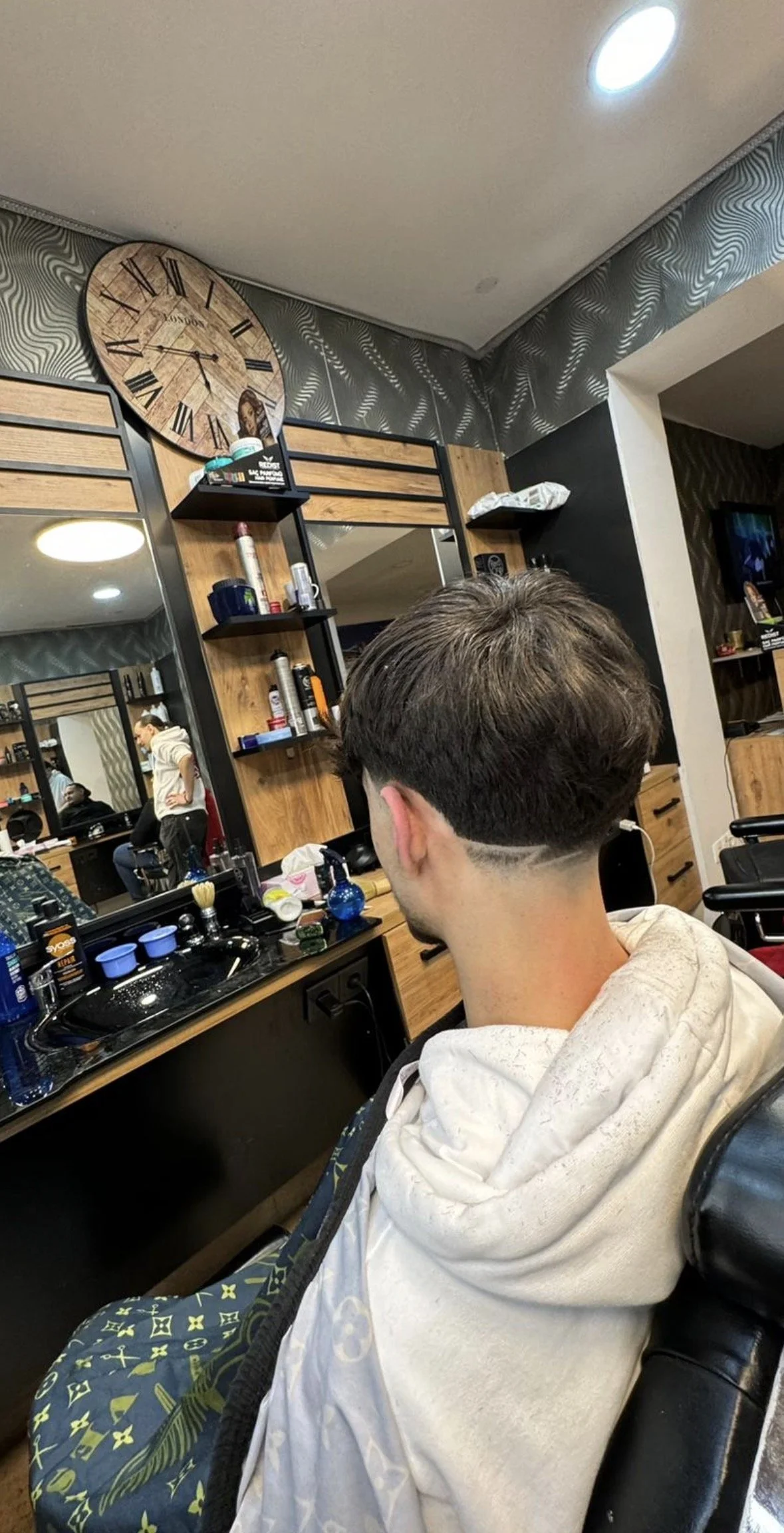A man with a dark, neatly styled haircut with a shaved line design on the back of his head, sitting in a barber's chair in a barbershop with a black and wood interior, a mirror, and grooming supplies on the counter.