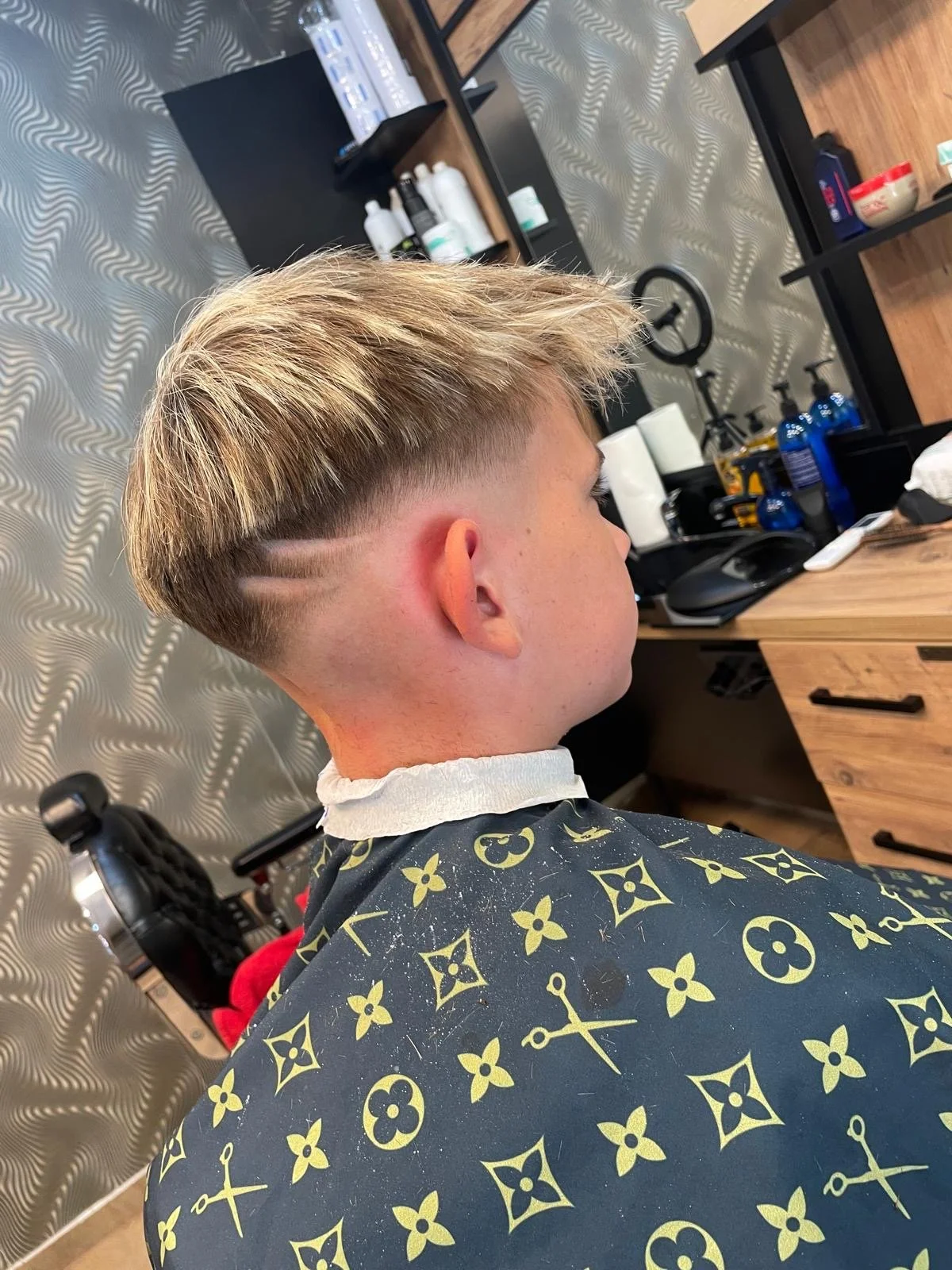 A person with a short, blond hairstyle featuring unique shaved lines on the side, sitting in a barbershop chair with a salon cape, facing a mirror and a workspace with hair products, tools, and a computer mouse.