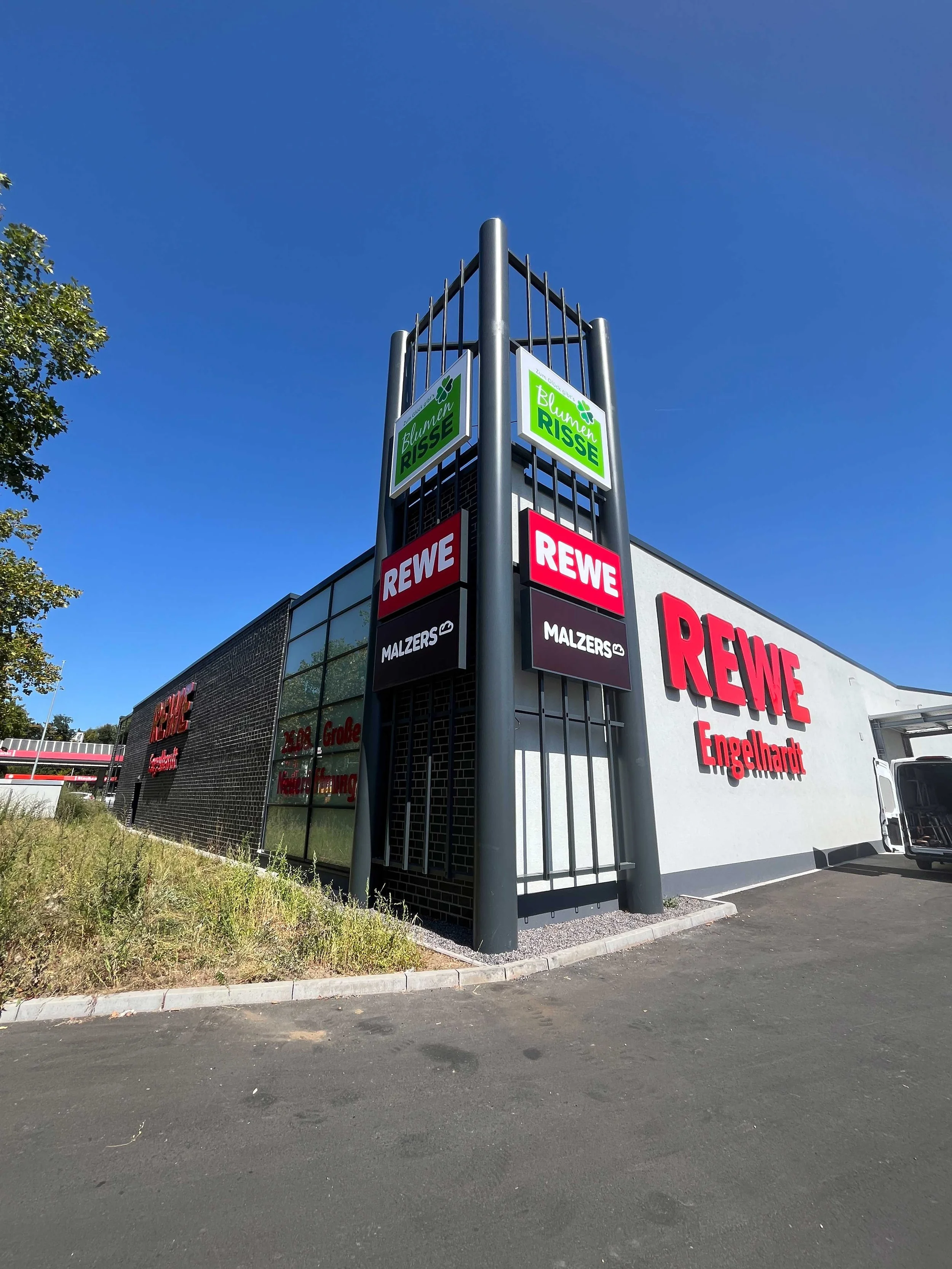 Außenansicht eines modernen Supermarktes mit Schildern von REWE, Malzers, Blumen Risse und Engelhardt auf einem grauen Gebäude mit dunklen Ziegelsteinen und einer Parkfläche im Vordergrund.