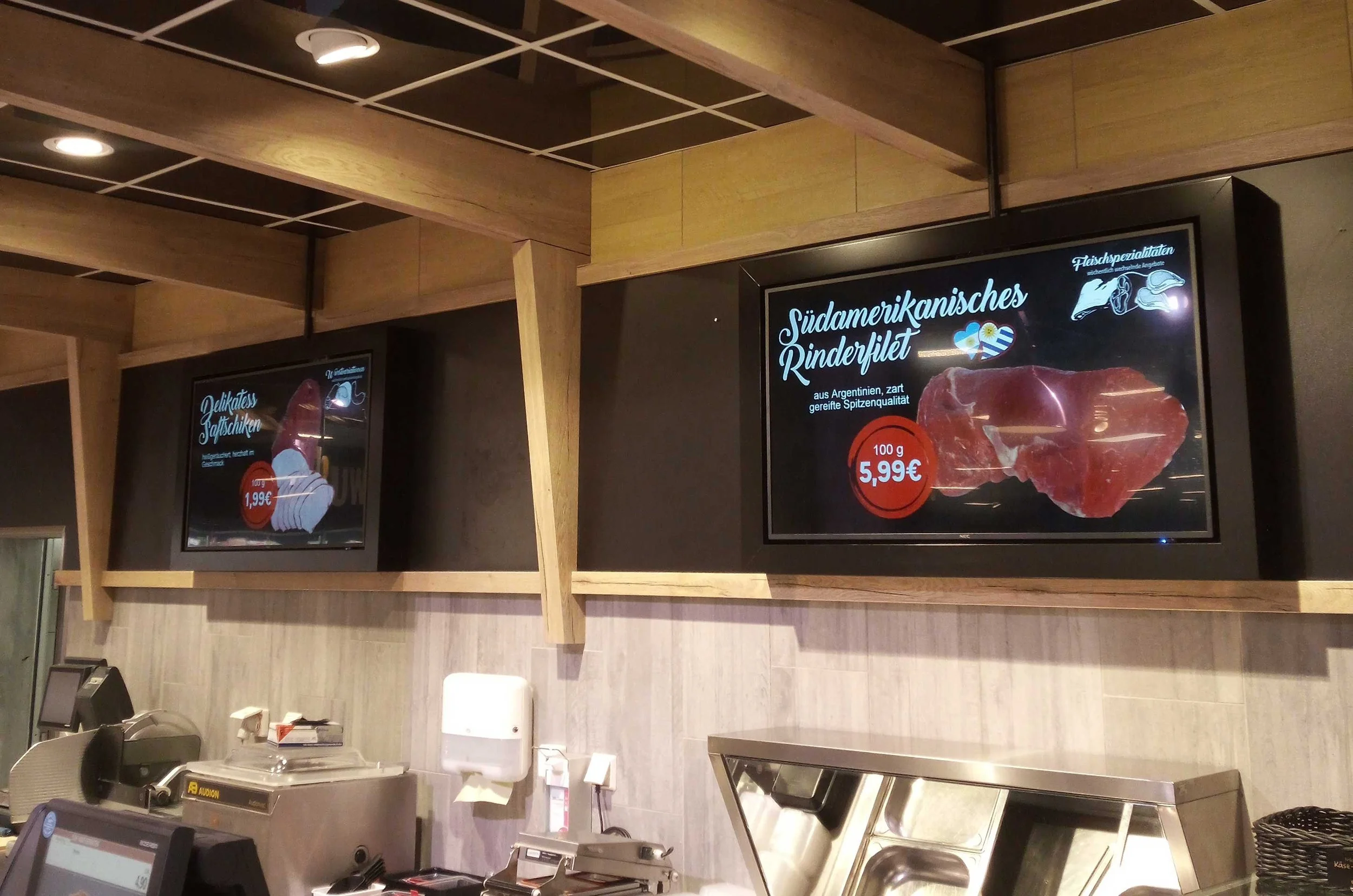 Digitale Menükarten im Nutzungsraum eines Supermarkts, die auf Fleischspezialitäten aus Argentinien hinweisen. Das rechte Display zeigt ein Stück Rindfleisch mit dem Preis von 5,99 € pro 100 g, die linke Anzeige bewirbt Delikatess-Jakobsmuscheln.