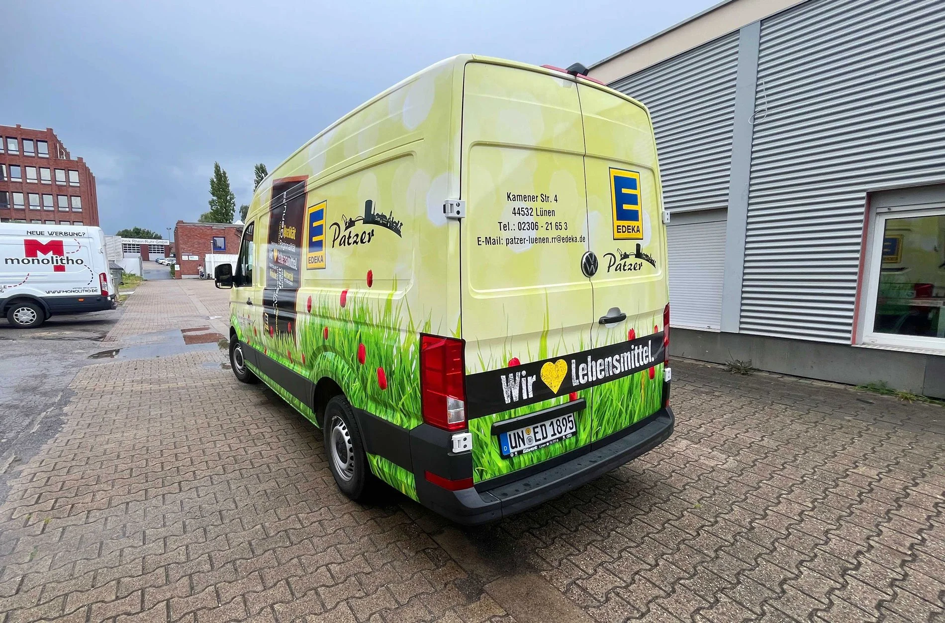 Ein gelber Lieferwagen mit Graskonzept, Edeka-Logo und Schriftzug "Wir ♥ Lebensmittel" steht vor einem Gebäude. Im Hintergrund sind weitere Fahrzeuge, eine rote Backstein-Gebäude und ein grauer Himmel zu sehen.
