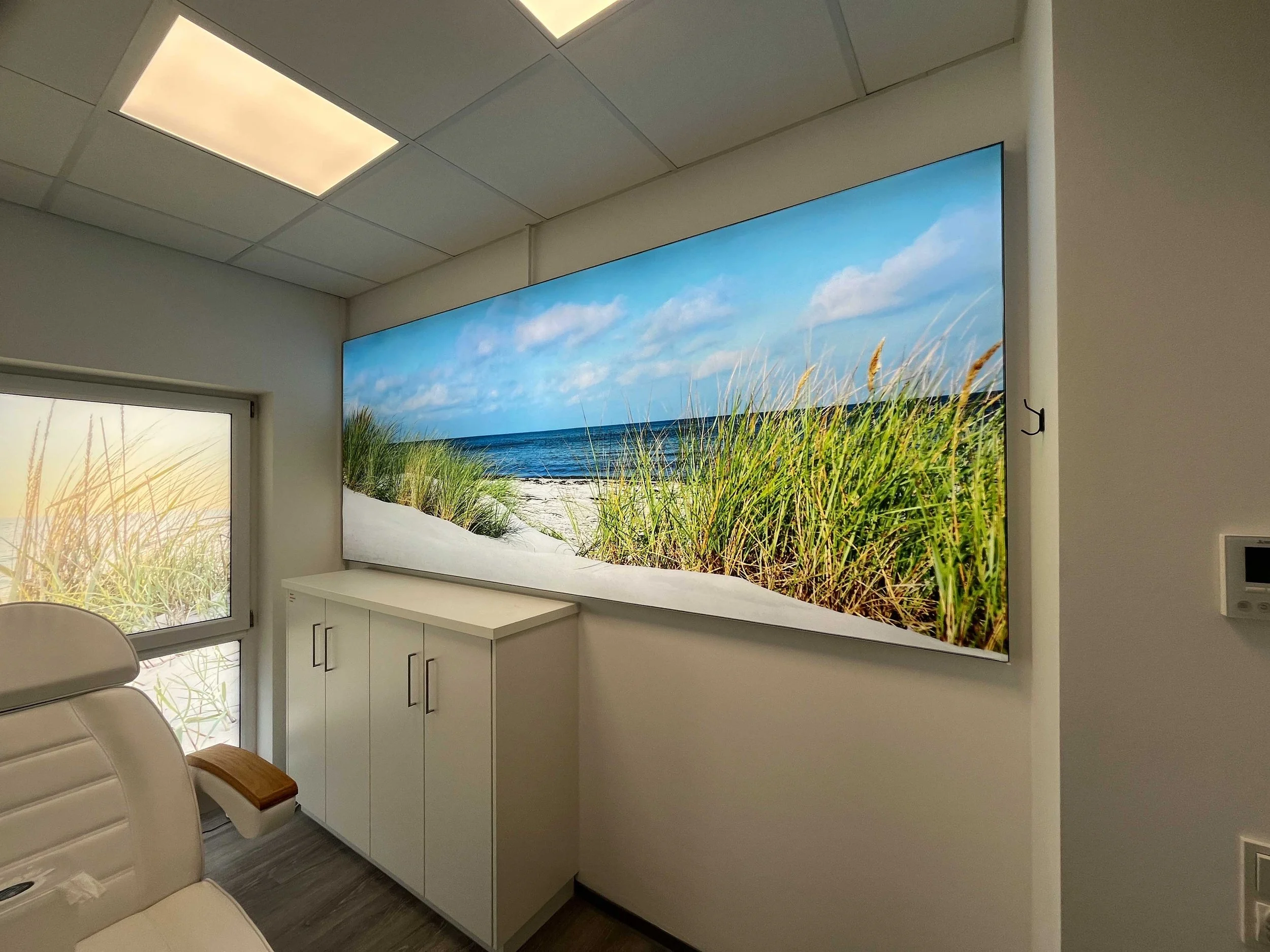Ein Raum mit einer großen Wandbildprojektion eines Strandes mit Blauen Himmel, Sand und Gräser, einem weißen Schrank und einigen weißen Stühlen.