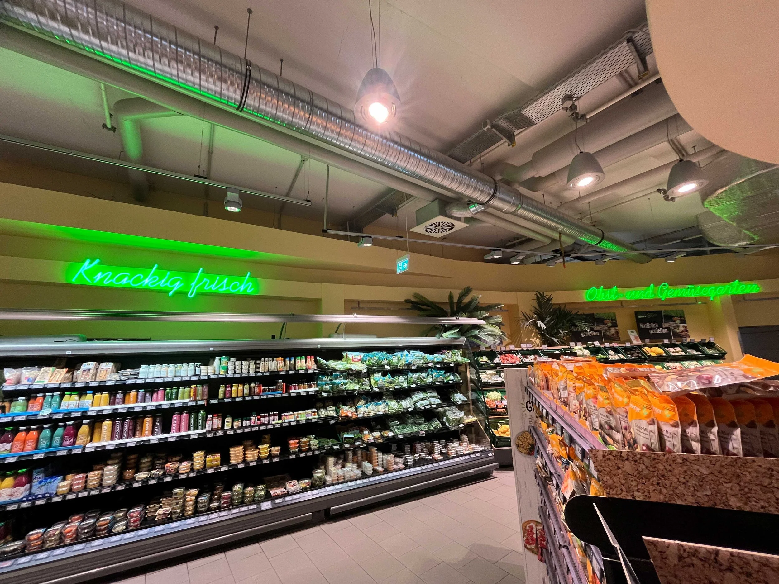 Blick in die Obst- und Gemüseabteilung eines Supermarkts mit Lebensmittelregalen, Obstkisten und einer neon-grünen Leuchtbeschriftung.