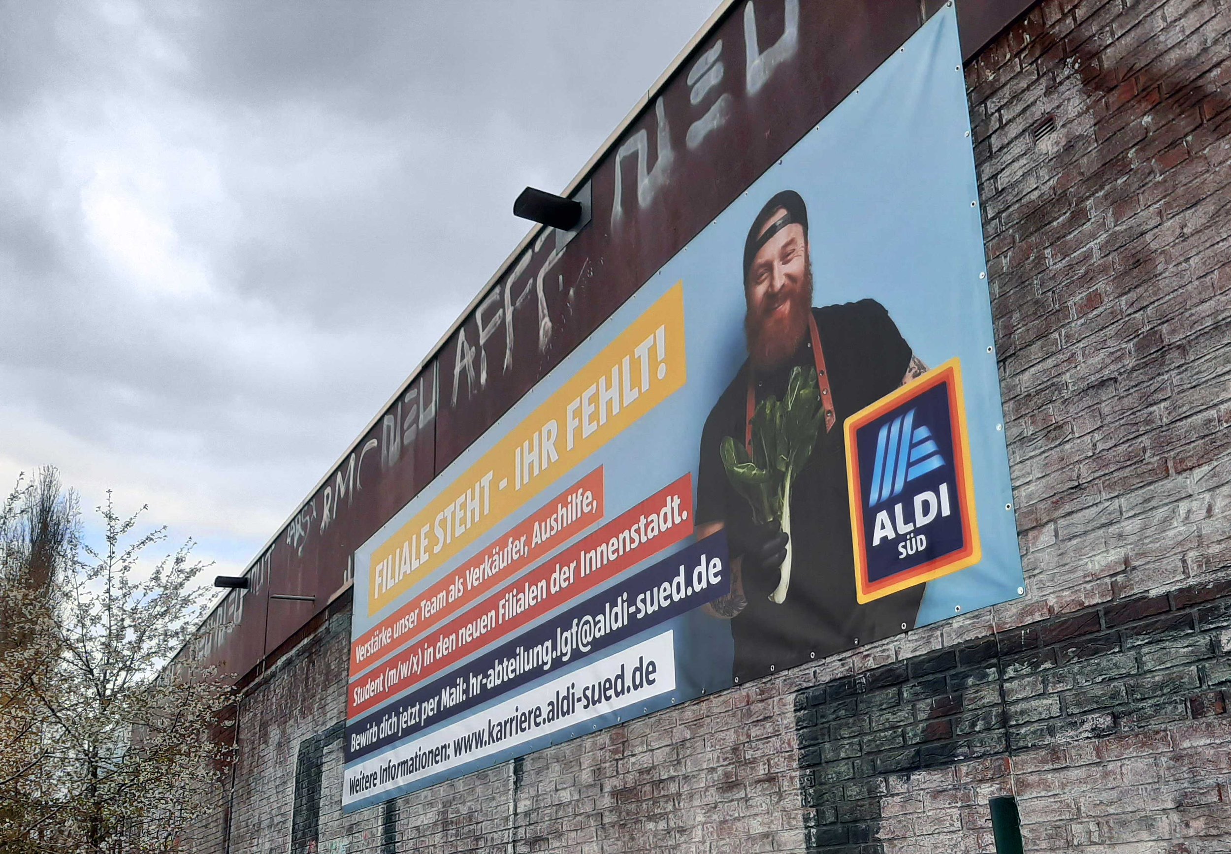 Werbebanner in deutscher Sprache mit einem lachenden Mann und Aldi Süd Logo, das auf eine Filialsuche und Stellenangebote bei Aldi Süd hinweist, an einer Ziegelmauer befestigt.