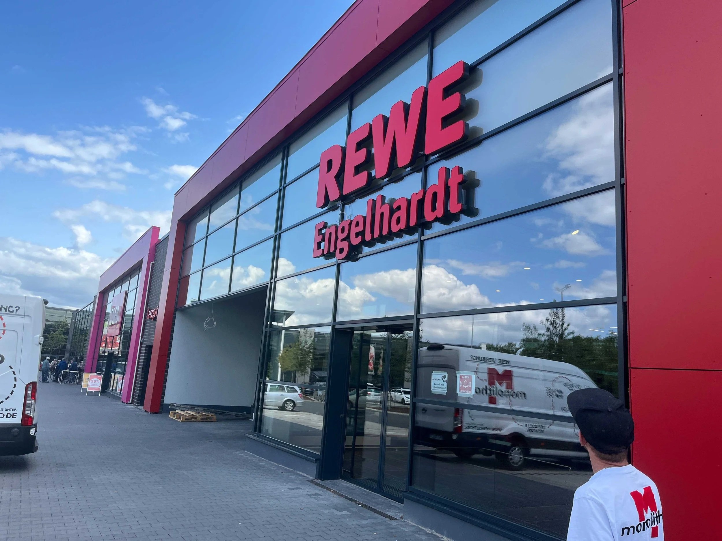 Außenansicht eines Gebäudes mit der Aufschrift 'REWE Engelhardt' auf der Front. Es sind Fahrzeuge im Parkplatz vor dem Geschäft zu sehen, und die große Glasfassade spiegelt den Himmel mit Wolken wider.