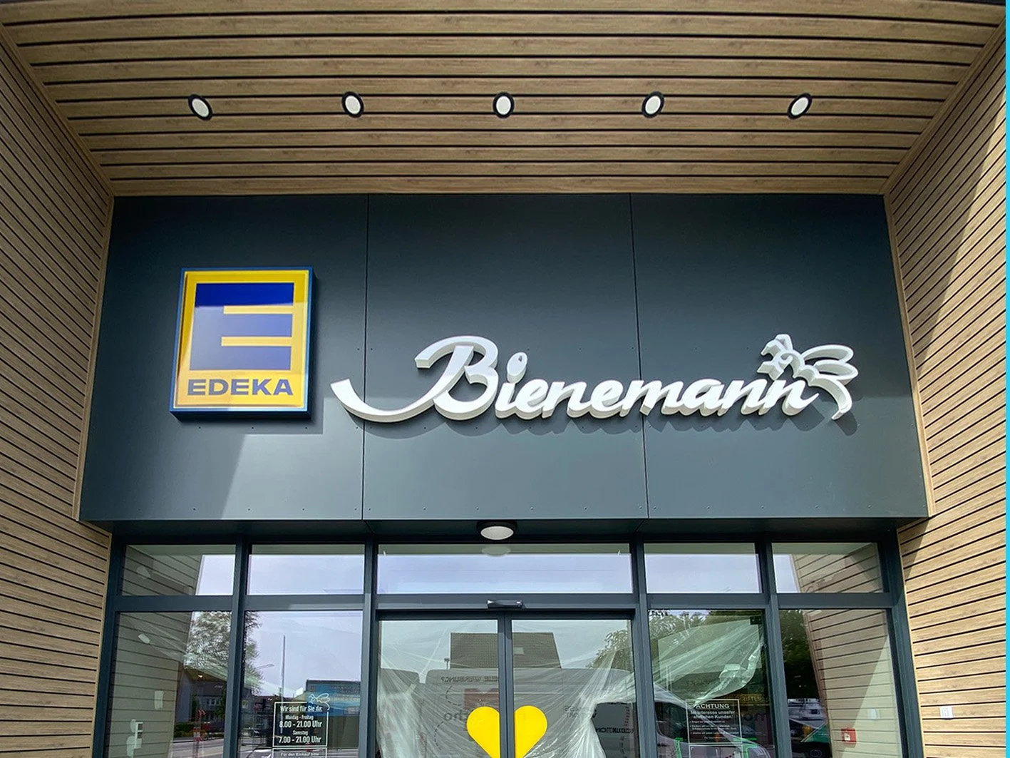 Vorderansicht eines Geschäftseingangs mit Edeka und Bienenmani-Schriftzug an der Fassade, umgeben von Holz- und Metallverkleidung.