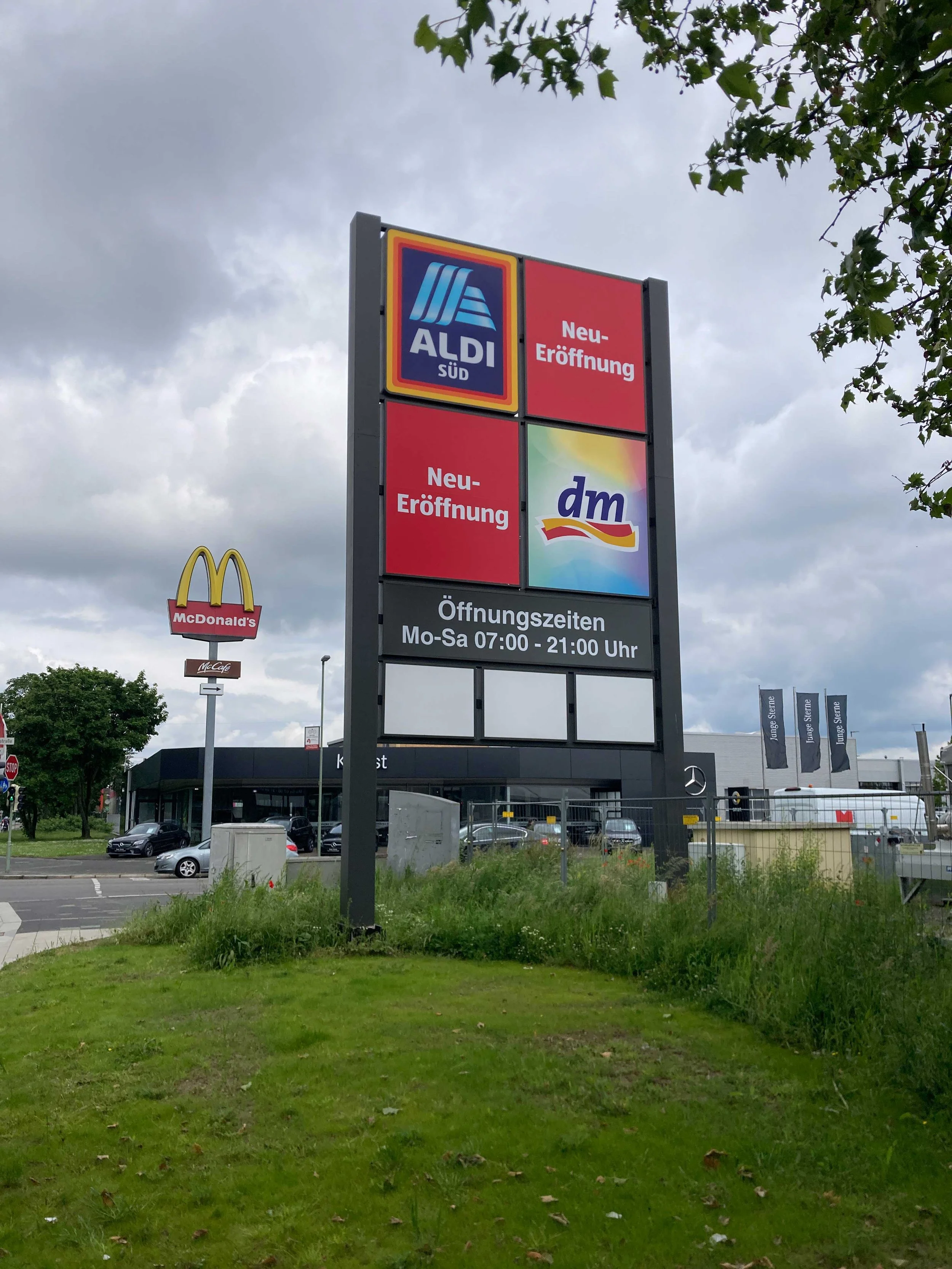 Werbetafel vor einem Supermarkt zeigt die Logos von Aldi Süd und dm, mit Angaben zu den Neueröffnungen und Öffnungszeiten. Im Hintergrund sind McDonald's und andere Geschäfte sichtbar, bei bewölktem Himmel.