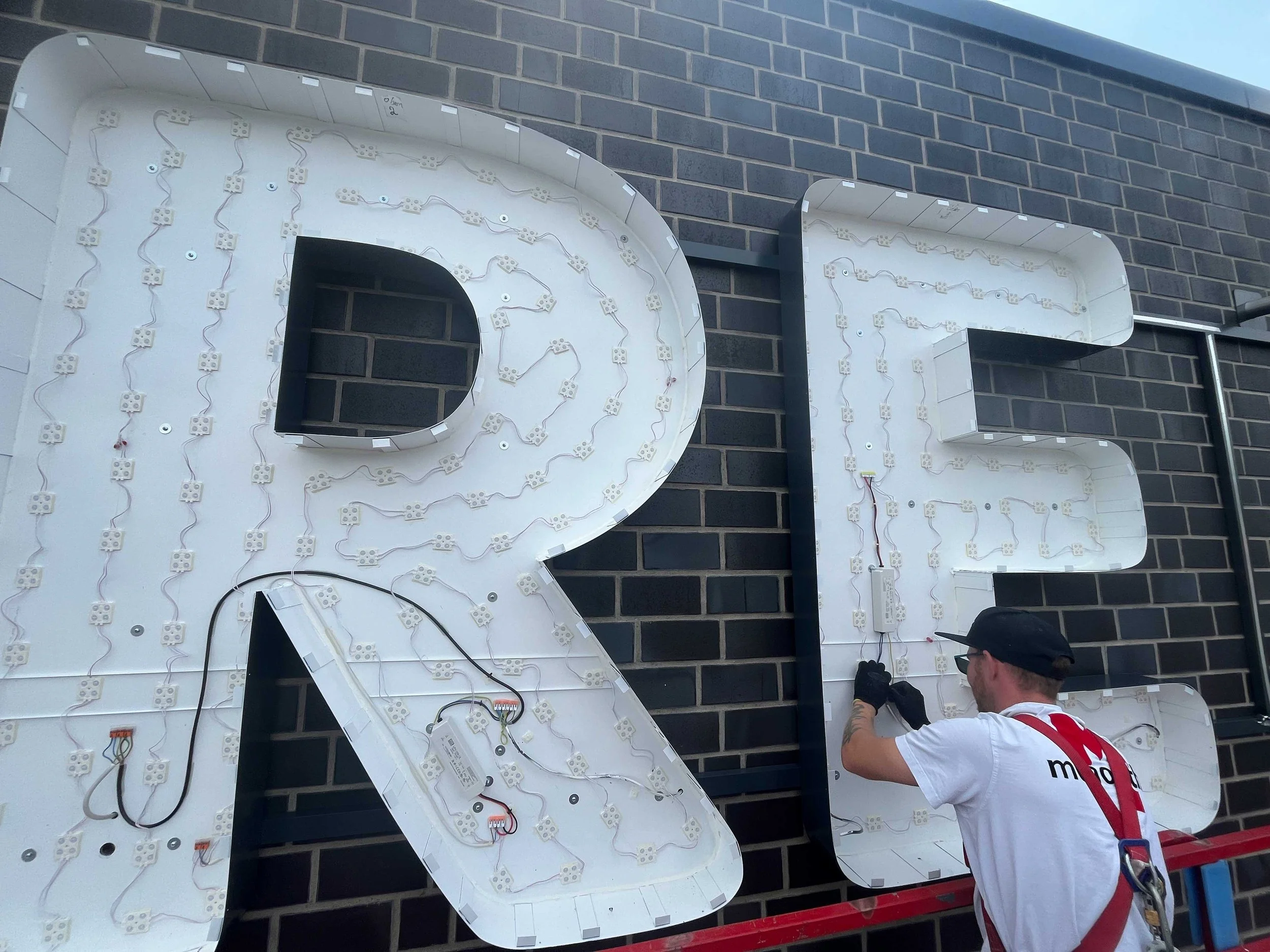 Ein Handwerker montiert eine große Leuchtreklame auf einer Außenwand aus Ziegelsteinen. Er arbeitet an der Rückseite der beleuchteten Buchstaben, die noch nicht vollständig fertiggestellt sind.
