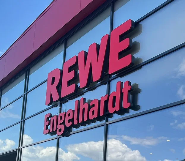 Außenansicht eines Gebäudes mit der Beschriftung "REWE Engelhardt" auf der Glasfassade, reflektierende Wolken und blauer Himmel