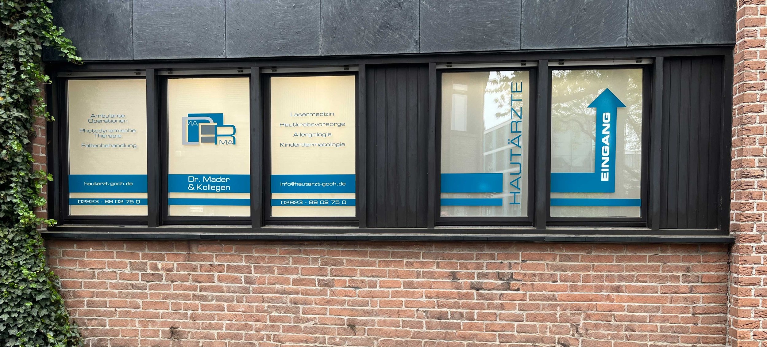 Fenster eines medizinischen Fachzentrums mit blauen und weißen Beschriftungen, die verschiedene medizinische Dienstleistungen wie Lasertherapie, Hautkrebs-Vorsorge und Kinderdermatologie anzeigen.