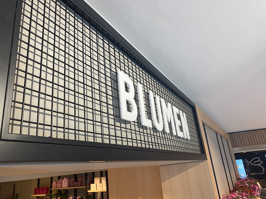 Das Bild zeigt ein Schild mit dem Wort 'BLUME' in weißen Buchstaben auf einer Gitterwand, vermutlich in einem Innenraum oder einer Verkaufsstelle.