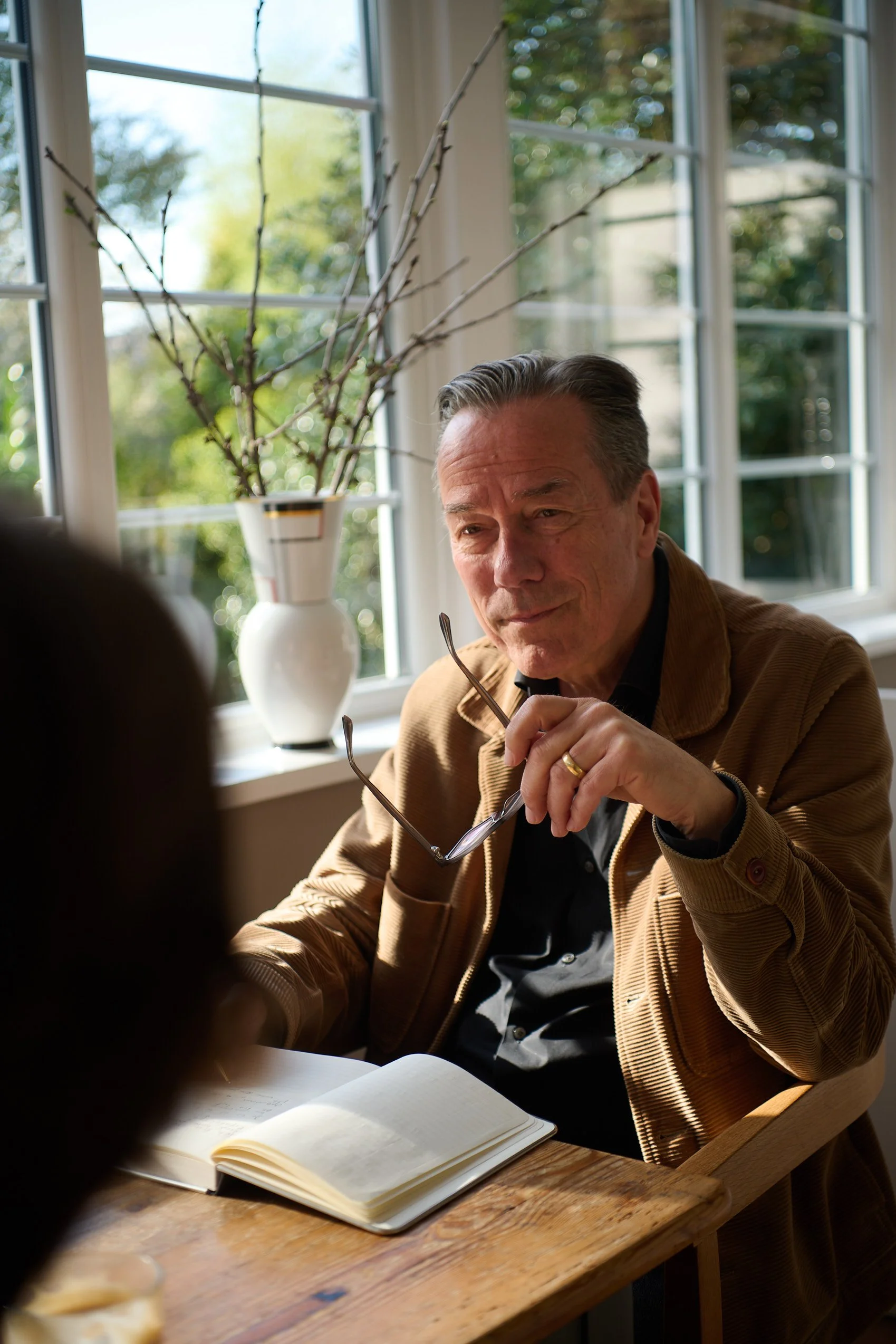 Christopher Gerisch trägt eine braune Jacke und sitzt an einem Holztisch mit einem offenen Notizbuch. Er hält eine Brille in der Hand und schaut nachdenklich. Das Gespräch über die verstorbene Person. Inhalte für die Trauerrede