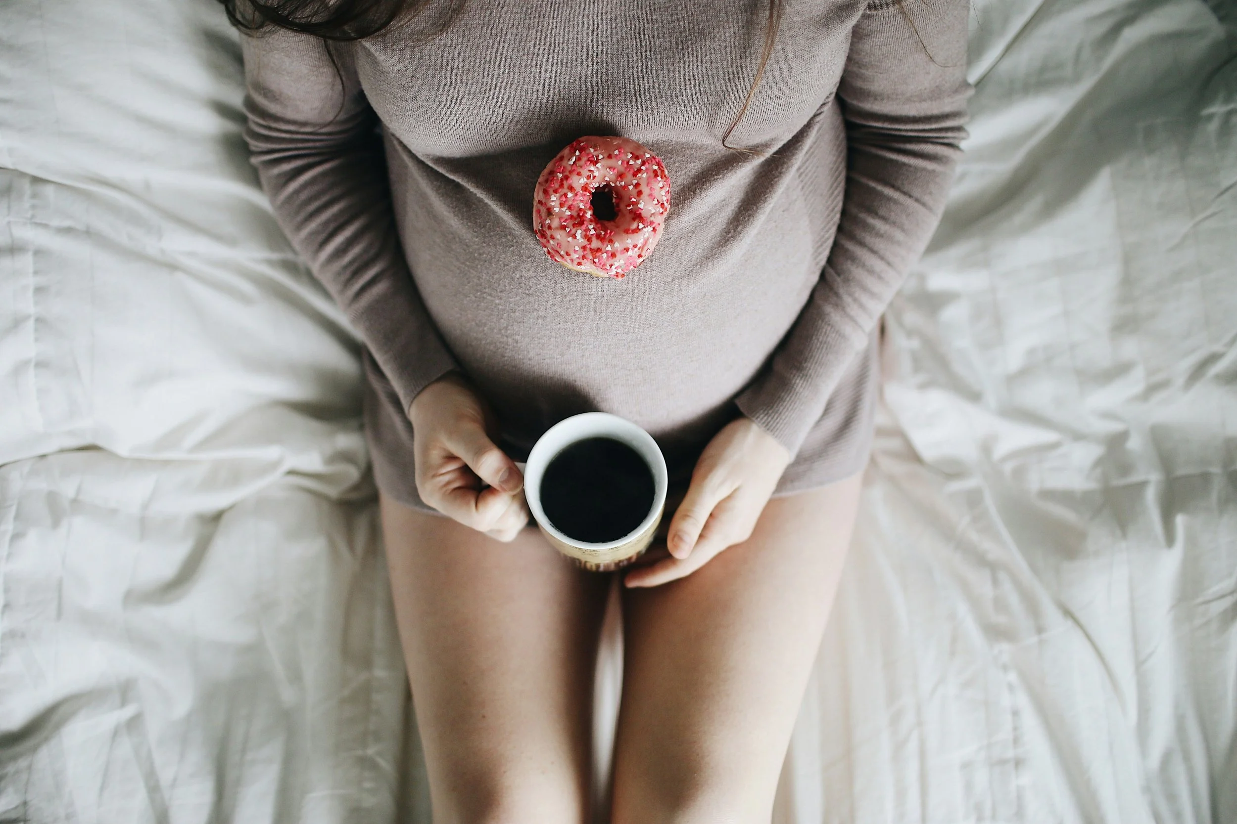 Pregnant woman in cozy sweater holding coffee mug with a donut resting on her belly representing maternal health